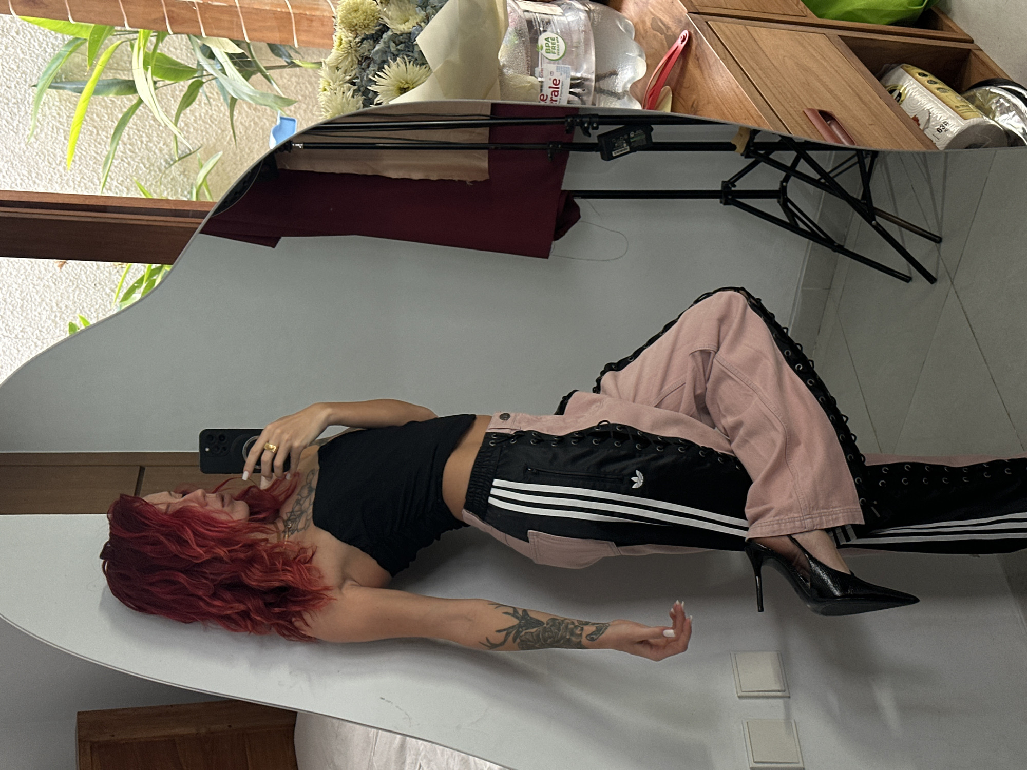 A woman with vibrant red wavy hair is leaning against a white wall in what appears to be a bedroom or dressing area. She is wearing a black strapless top and pink pants with black Adidas stripes and lace-up details. She is holding a black smartphone in her right hand, taking a selfie in a large, irregularly shaped mirror that reflects the room. Her left arm is resting on the wall, showcasing a tattoo on her forearm. Her left leg is bent at the knee, with her foot in a black pointed-toe high heel.
The mirror also reflects a wooden dresser with drawers and shelves, on which sits a bouquet of flowers in a clear plastic bottle of water. Behind the flowers, a large window shows lush green plants and possibly a patio area. A tripod is also visible in the reflection. The overall environment suggests a stylish, possibly bohemian, personal space. The presence of a large mirror and the way the woman is posed indicate she is capturing a fashion or lifestyle moment.