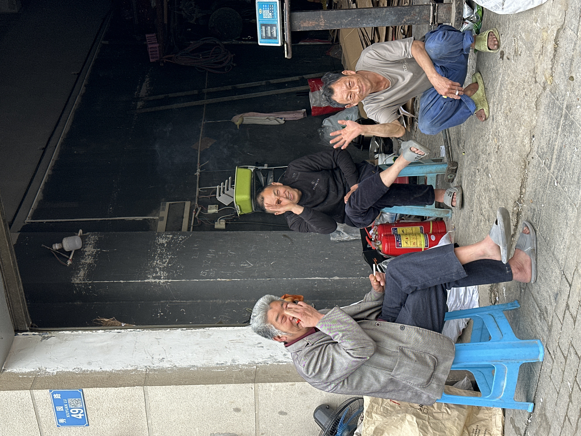 This media file captures three men taking a break on a sidewalk, appearing to be in good spirits, likely in Jiangjin, China.
In the foreground, an older man with grey hair and a grey jacket is seated on a bright blue plastic stool, smiling broadly with his hand near his face, possibly holding a cigarette or small item to his mouth. To his right, a red fire extinguisher stands on the ground. Next to him, a man in a black hoodie and dark pants sits on another blue plastic stool, laughing with his hand covering part of his face. Further back and to the right, a third man in a light t-shirt and blue pants is squatting, holding a lit cigarette and gesturing with his left hand, also smiling. All three men are wearing casual slippers.
The setting appears to be outside a dark storefront or workshop, indicated by the obscured interior in the background which contains wires, equipment, and a hanging digital weighing scale. The ground is paved concrete.
Notable details include:
* **Emotions:** All three men are clearly smiling or laughing, conveying a sense of camaraderie and enjoyment.
* **Time of day:** The bright, even lighting suggests daytime.
* **Location cues:** A blue street sign in the bottom left corner reads "秀园路 XIUYUAN LU 49", indicating Xiuyuan Road, No. 49. It also features a QR code.
* **Visible text:** Besides the street sign, the digital scale shows "3-2020", "江街道" (Jiangjiedao), and various numbers like "2215-7", "25,32:14.5", "26521". The man in the black hoodie has text on his sleeve, partially visible as "AR DEBBUBR RO".
The men are engaged in a relaxed interaction, possibly sharing a lighthearted moment during their break.