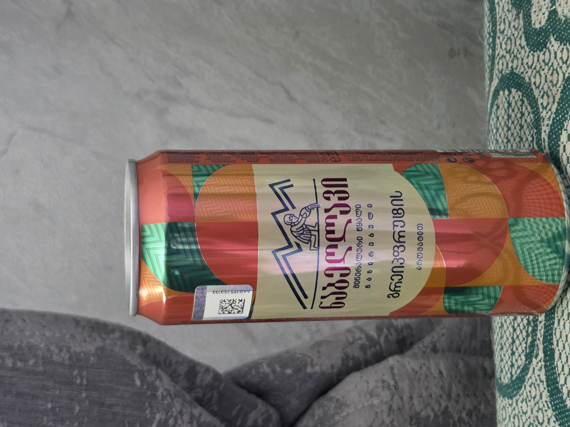 A close-up shot of a can of "Nabeghlavi" mineral water with grapefruit flavor. The can has a distinctive design with geometric patterns in green, orange, and red. The brand name and description are written in Georgian script. A barcode and number "AAB355350100" are visible on the side. The can is positioned on a surface with a grey and green patterned textile.