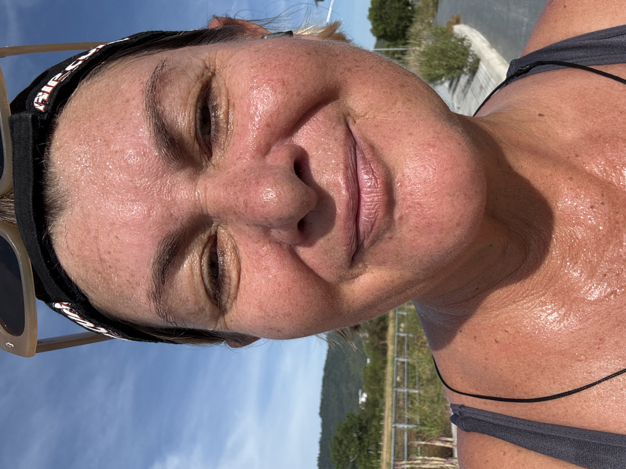 A close-up, slightly rotated selfie depicts a woman with fair skin and sweat on her face and chest. She is wearing a black headband and light-framed sunglasses are visible to the left. Her eyes are partially closed in a smile. Behind her, the outdoor scene includes a fence, some greenery, and a distant mountain range under a blue sky. Based on the prompt, the location is Baan Chaweng, Thailand.