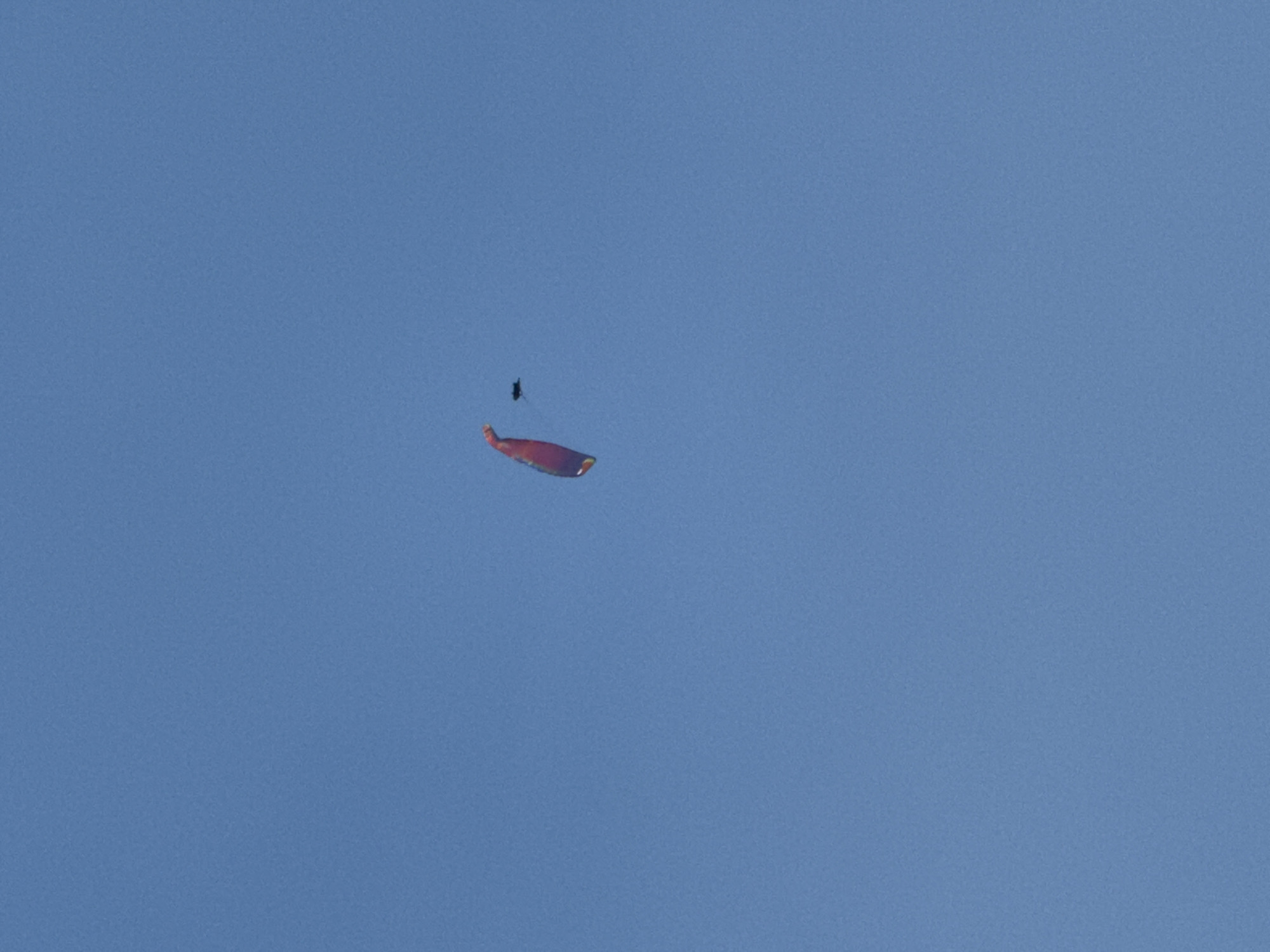 A colorful kite, predominantly red and yellow, is in flight against a clear blue sky. To the upper right of the kite, a small dark object, possibly a drone or a bird, is also visible. The image was captured in Boreti, Montenegro, during daylight hours.