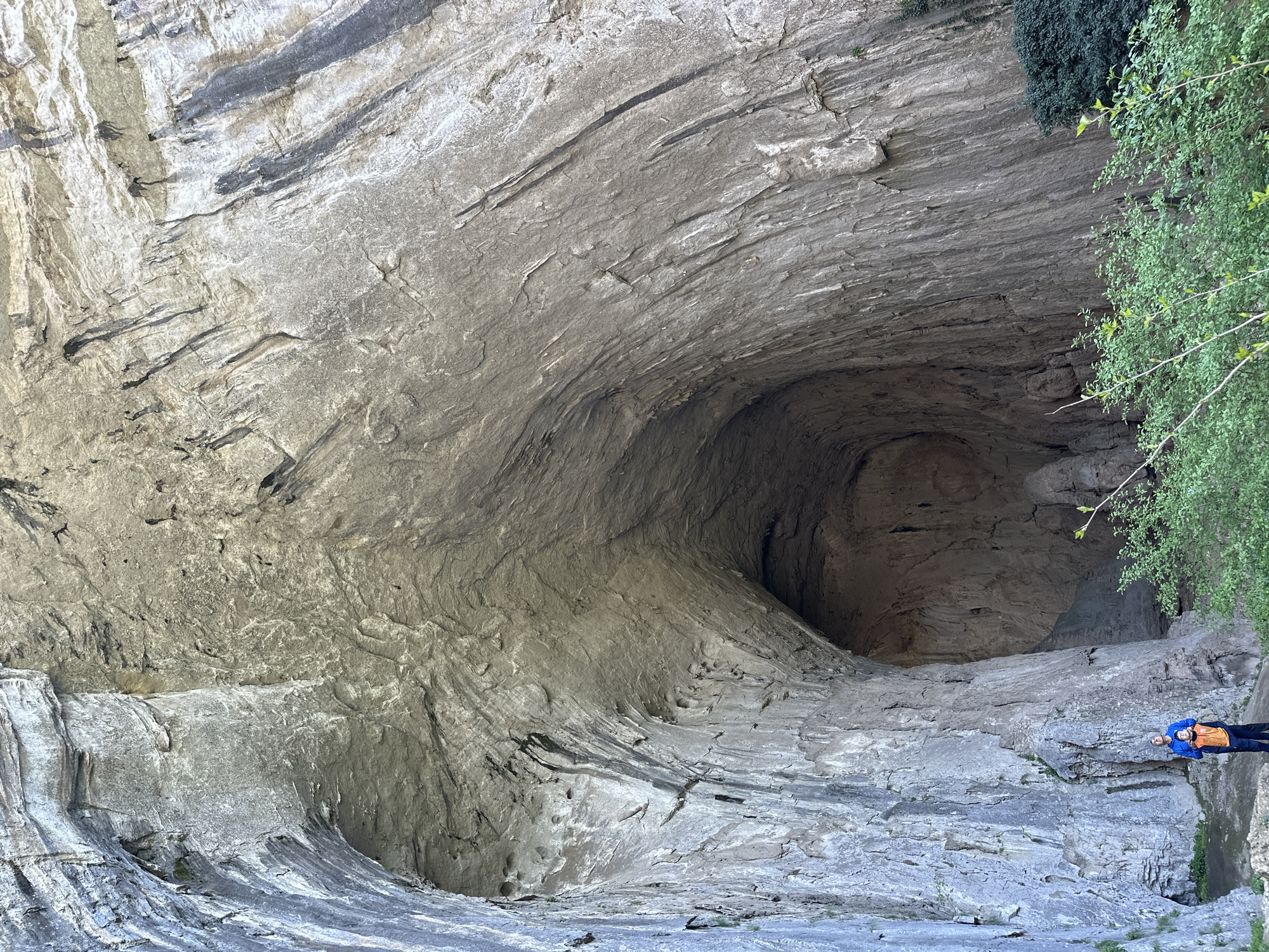 The image shows a large cave entrance in a rock formation. The cave is located in the city of Karlukovo, Bulgaria. The rock is gray and layered. The cave entrance is dark. On the right side of the image, a person wearing a blue jacket and an orange vest is standing near the edge of the formation. Green foliage is visible near the person.