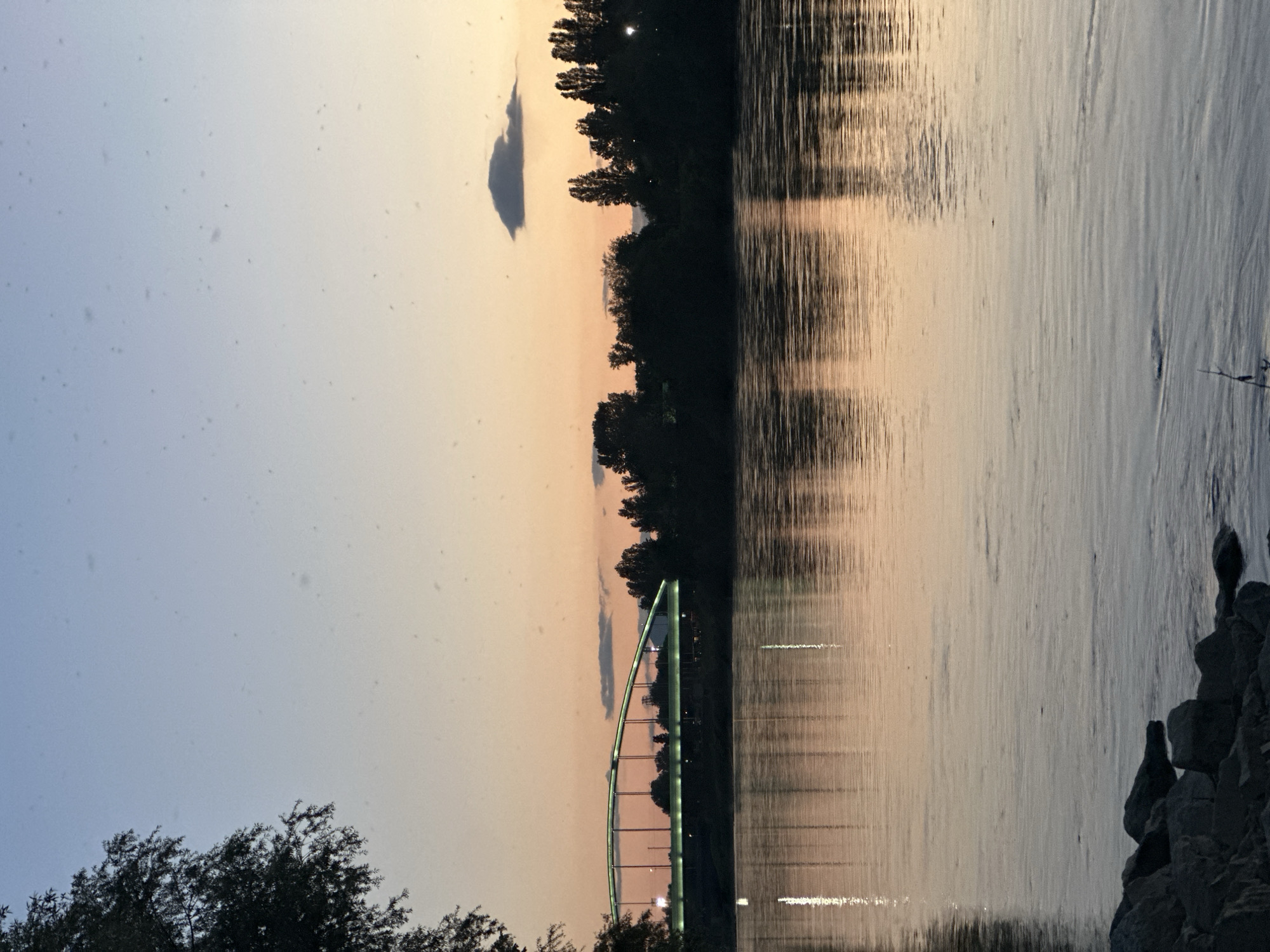 The image shows a body of water with the reflection of trees and sky on its surface. A bridge with green lighting is visible in the distance. The sky transitions from light blue to orange. There are trees on the opposite bank and rocks on the near bank. The image was taken in Zagreb, Croatia.