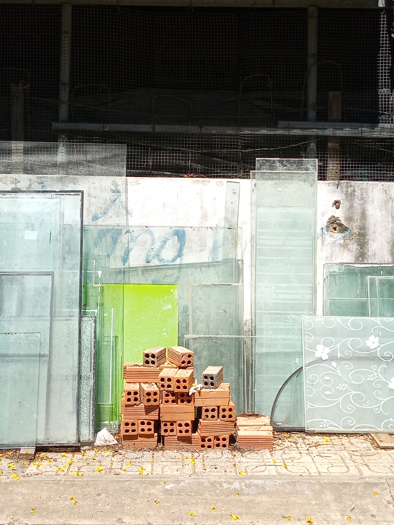 The image depicts an outdoor, street-level scene in Nha Trang, Vietnam. In the foreground, a concrete sidewalk, composed of large square tiles, is littered with numerous yellow petals. Two uneven stacks of red, perforated clay bricks are positioned on the sidewalk, with a small white plastic bag visible near the base of one stack.
Immediately behind these objects, numerous sheets of glass are propped vertically against a concrete wall. These glass panels vary in size and transparency; some are clear, others are frosted, and several feature etched designs, including floral patterns on a lower right panel and bamboo motifs on a taller central panel. A prominent, rectangular lime-green panel or door is also visible among the glass sheets.
The concrete wall behind the glass is weathered, showing discoloration and faded blue graffiti. Above this wall, a dark, gridded mesh structure, resembling a security screen or enclosure, occupies the upper half of the image. Within this meshed area, dark support columns and tangled wires are discernible. No individuals are present in the scene, and no actions or interactions are depicted. The lighting indicates direct sunlight.