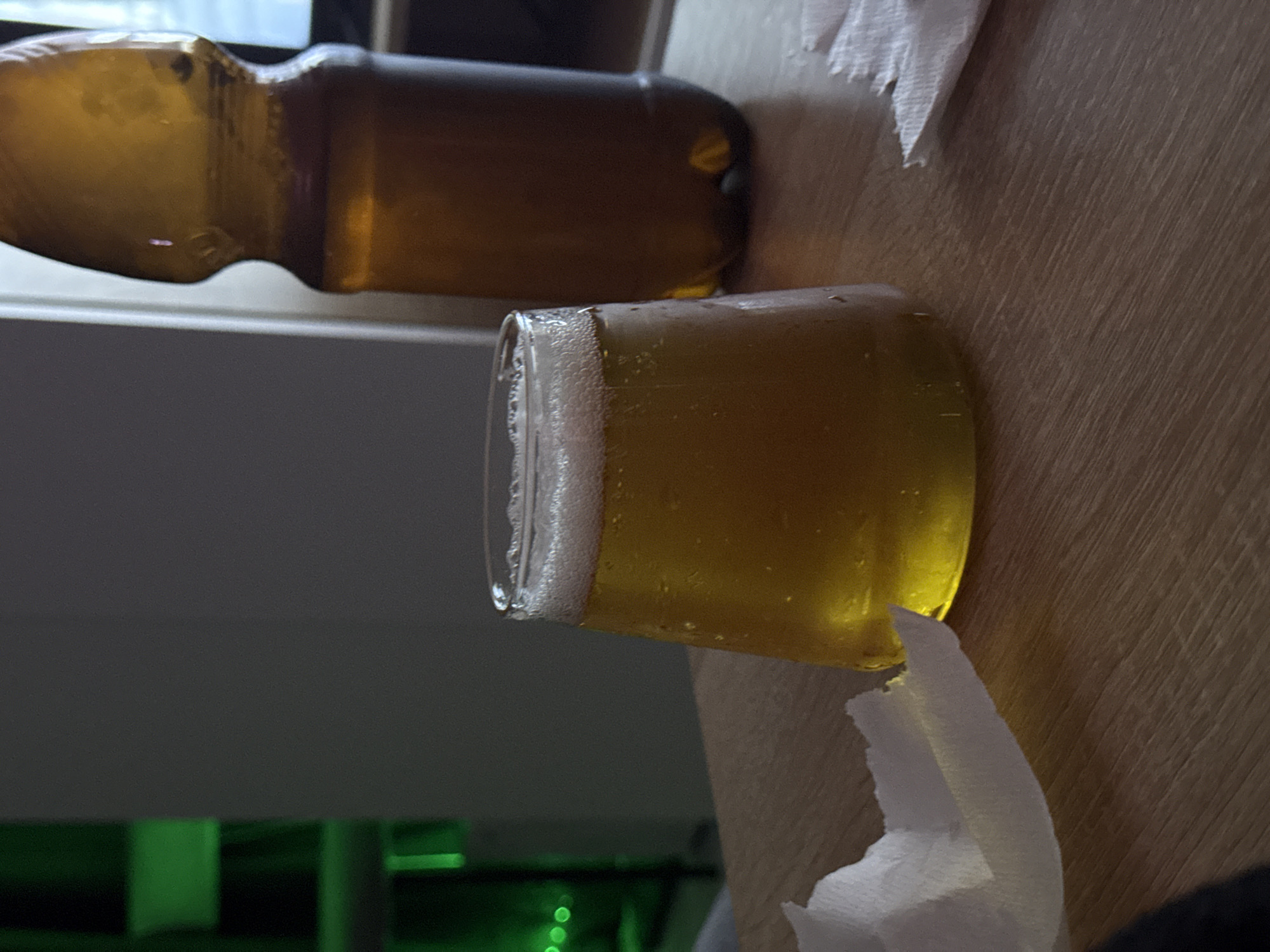 A glass of beer with a foamy head sits on a wooden table alongside a partially visible beer bottle and two white napkins. The background reveals a dark wall with some green lights visible at the bottom. The location is unknown.