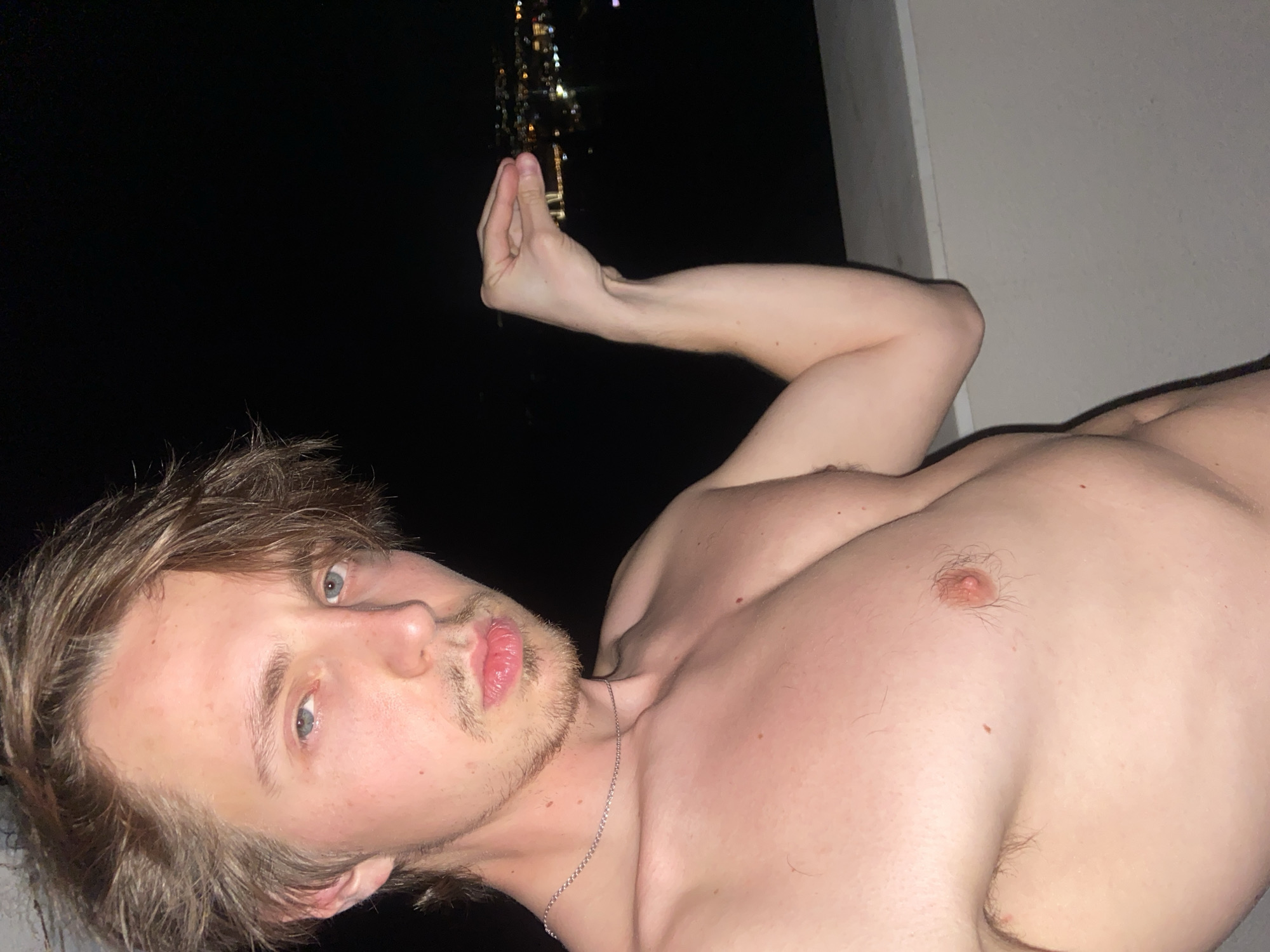 A shirtless, light-skinned male with light brown hair and blue eyes lies on his side. He has a light beard. He is wearing a silver chain around his neck. His left arm is raised and his hand is in a pinched gesture. In the background, blurry lights indicate a cityscape at night. The photo appears to be taken outdoors, near a building structure. There is no clear indication the location is Šušanj, Montenegro.