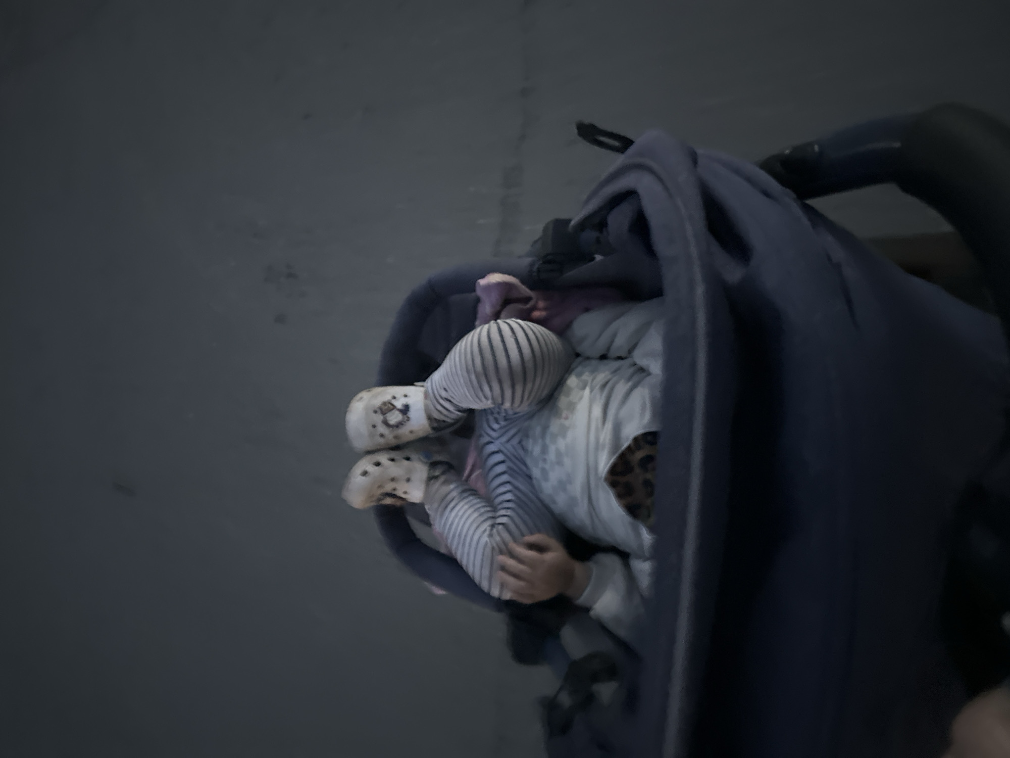 A low-angle, blurry shot depicts a young child upside down in a dark blue stroller. The child's feet, clad in white Crocs with a pattern, are pointed upwards, with their legs bent at the knees. They are wearing striped pants and a long-sleeved shirt with a leopard print detail on the chest. One of the child's hands is visible. The stroller is on a paved surface, likely a sidewalk or road, in Chaikovsky, Russia. The background is a blurred, greyish expanse, suggesting an outdoor environment. The overall lighting is dim, and the image appears to have been taken in motion, contributing to the blurriness.