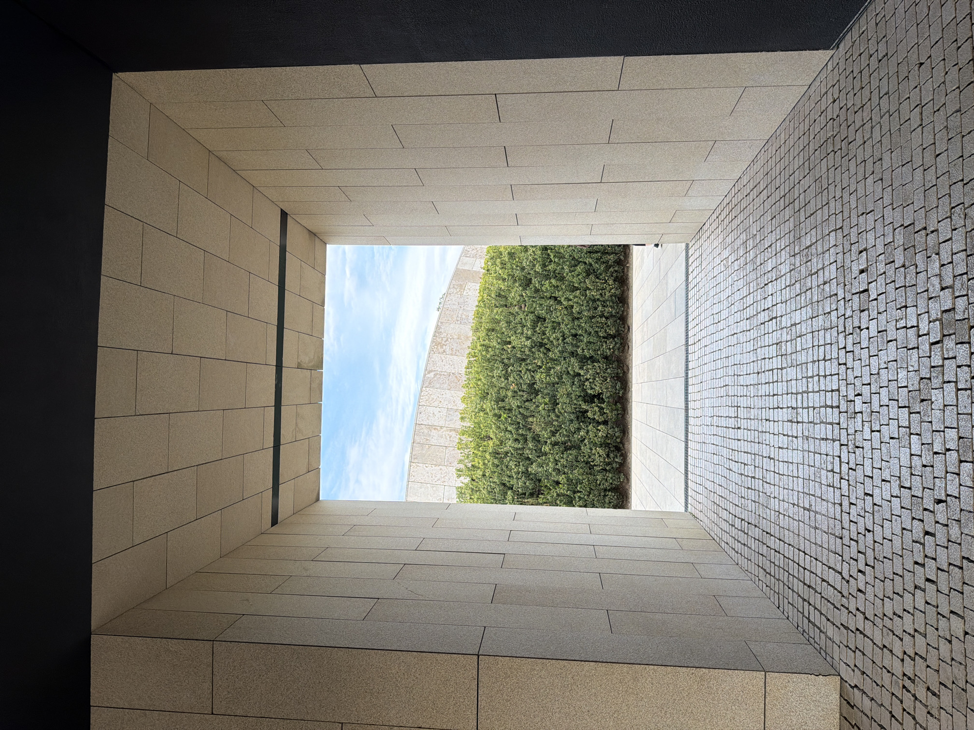 The image shows a view from within a narrow, rectangular opening or tunnel-like structure. The left and bottom walls are constructed from large, light-colored stone blocks arranged in a linear pattern. The right wall is composed of small, gray cobblestones. A black, flat surface forms the top of the opening, partially obscuring the view.
Looking through the opening, a clear blue sky with light clouds is visible above a tall, thick hedge of green bushes. A curved, light-colored wall runs along the left side of the hedge.
There are no people visible in the image. The location is likely outdoors, as suggested by the sky and vegetation. It has been identified as the City of Krasnodar, Russia.