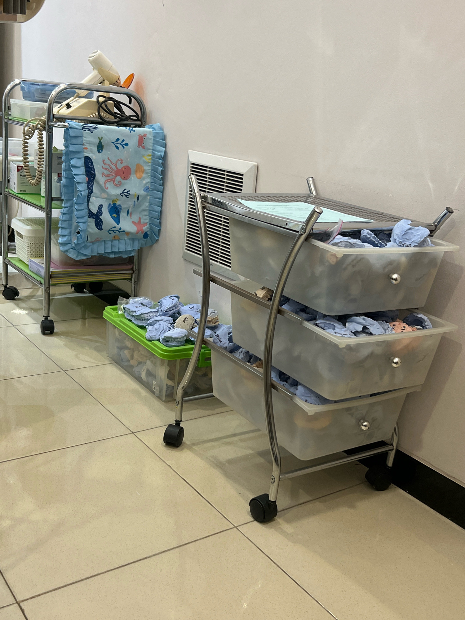 A medical examination room in Magelang, Indonesia, shows a multi-tiered chrome cart holding various medical supplies. The cart's shelves contain boxes of gauze pads, a blue blanket with an ocean-themed print, and a hairdryer. Below this, a green plastic bin is filled with what appear to be dental molds and blue surgical gloves. A second chrome cart with three translucent drawers is positioned beside it, each drawer containing more dental molds and blue gloves. The room has beige tiled flooring and a light-colored wall with a white air vent.