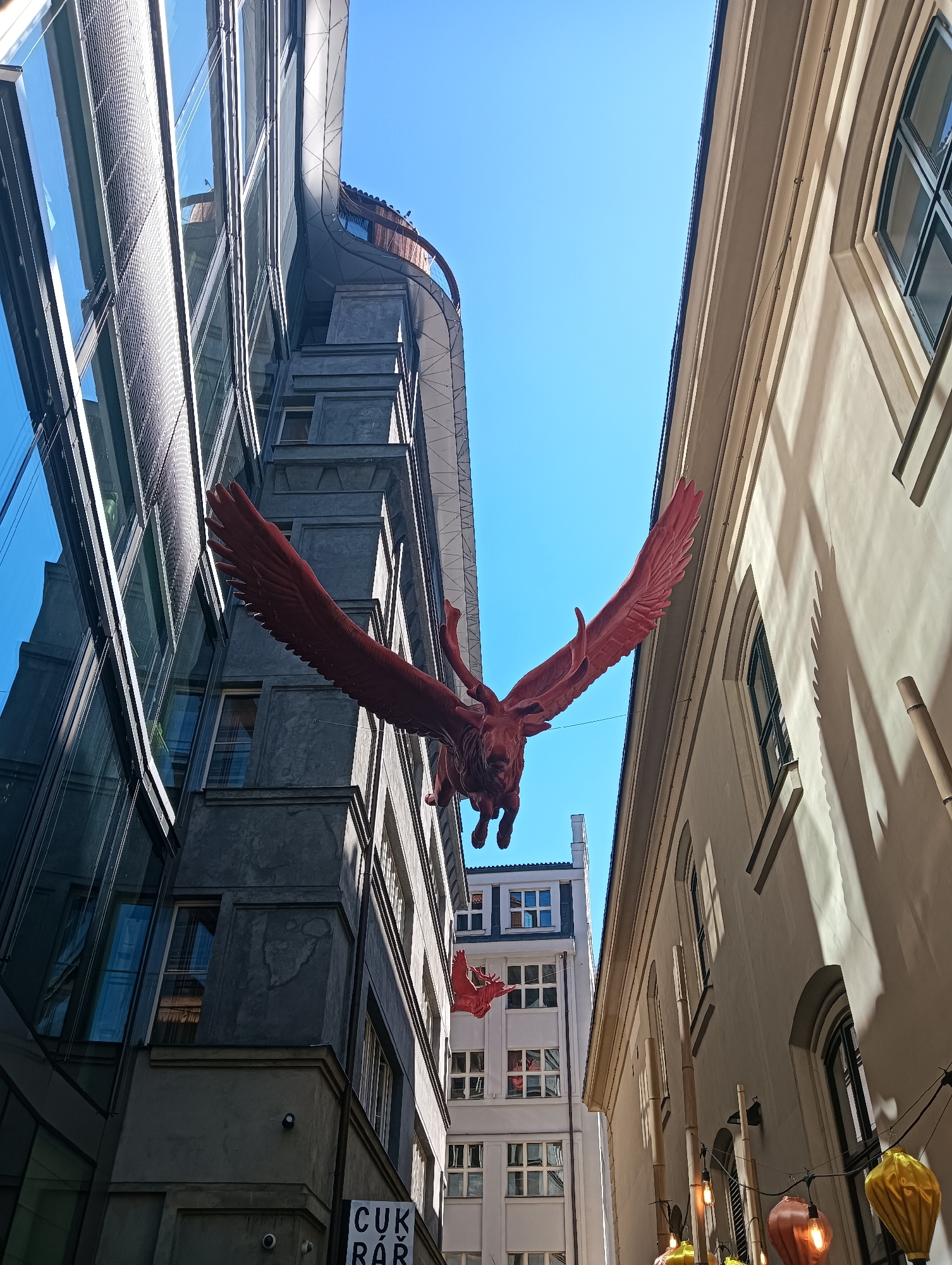 The image captures an upward view of a narrow street in Prague, Czechia, illuminated by a bright, clear blue sky. The scene is dominated by two contrasting buildings: on the left, a modern structure with a dark, reflective glass facade featuring a fine mesh pattern; on the right, an older, lighter-colored building with arched windows and decorative architectural elements. Suspended between the buildings, at a significant height, are two large, bright red sculptures of winged creatures, likely griffins or similar mythological beasts. The larger sculpture, prominently displayed, has its wings spread wide. A smaller, identical sculpture is visible further in the background. The street level is partially visible, showing elements of both buildings and a sign that reads "CUKR BAR" on the lower part of the modern building. Strings of colorful lanterns, one yellow and one reddish-brown, hang in the foreground on the right, adding a touch of festivity to the urban landscape. Shadows cast by the older building stretch across its facade, indicating the direction of the sun. The perspective is from ground level looking up, emphasizing the verticality of the architecture and the dramatic placement of the sculptures.
