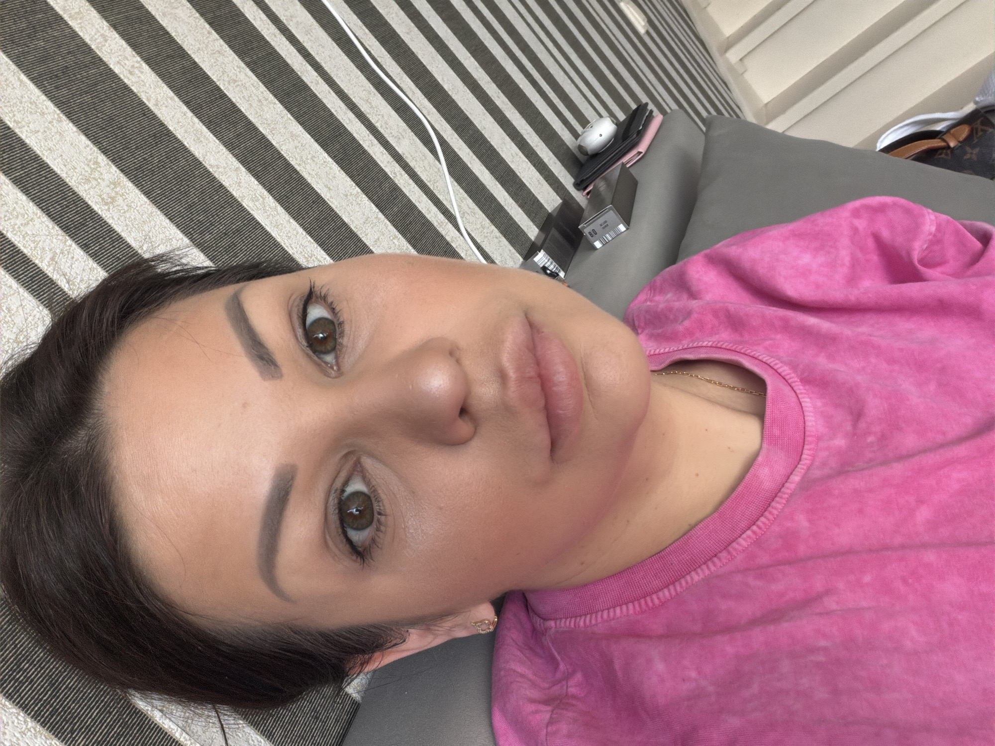 A close-up selfie shows a woman from the chest up, with her head turned to the right. She has brown hair pulled back, brown eyes, and is wearing a bright pink tie-dye t-shirt and a delicate gold necklace. She is wearing subtle makeup, including eyeliner and blush. Her left earlobe has a small gold earring. She is positioned in an indoor setting, possibly a living room, with a gray sofa visible on the left and a wall with a black and white striped pattern behind her. A white cord, possibly for electronics, runs diagonally across the striped background. To the right, on the striped surface, are a few small objects, including what appears to be a phone or tablet case and a small white electronic device. The lighting suggests it is daytime. There is no indication that this image was taken in Sochi, Russia.