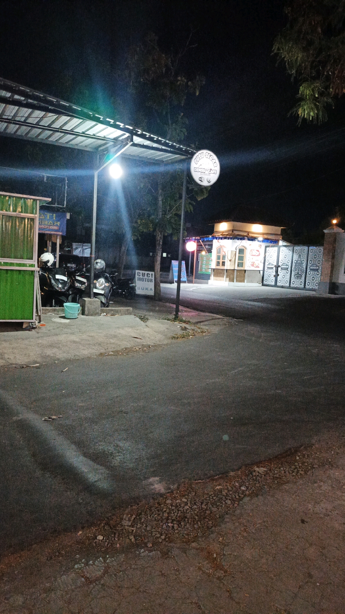 This nighttime image captures a street scene in Pabelan, Indonesia. A prominent circular sign with the text "SIRU SEGAR DENSUI CAFE" is visible, illuminated by a nearby lamp. To the left, a green corrugated metal structure partially obscures motorcycles parked on a concrete patch. A blue bucket sits on the ground near the motorcycles. Further down the street, a small building with a brightly lit interior and windows is situated behind a gate with an ornate, decorative pattern. A sign that reads "CUCI MOTOR BUKA" is also visible on a pole. The asphalt road is dark, with some debris and patches of gravel along its edge. Trees with dark foliage are present in the background, and the overall lighting suggests an urban or semi-urban setting at night.