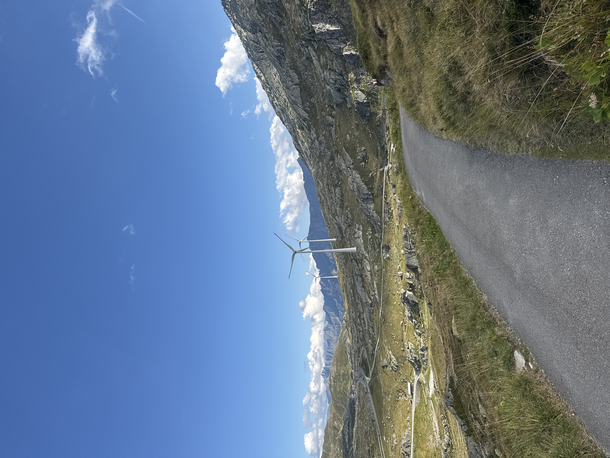The image captures a scenic view of the Gotthard Pass near Airolo, Switzerland, on a clear day with a vibrant blue sky dotted with white clouds. In the foreground, a paved road curves through a landscape of rolling hills covered in dry grass and scattered rocks. The road appears to be a walking or cycling path, with a person in red and white visible on the right side of the image.
In the mid-ground, a winding road snakes through the mountainous terrain, leading towards a cluster of buildings in the distance. Three prominent wind turbines stand tall against the backdrop of majestic, rocky mountains, their white structures contrasting with the natural landscape. The mountains themselves are characterized by rugged slopes and rocky outcrops. The overall environment suggests a remote, high-altitude location, typical of the Swiss Alps.
