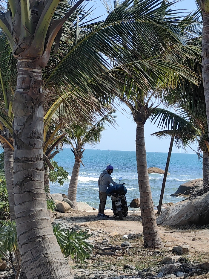 A man wearing a purple helmet stands near a motorbike laden with luggage, next to a beach. The ocean is visible in the background with some small boats out in the water. The scene is framed by multiple palm trees of varying sizes and distances, giving the impression of a tranquil tropical getaway. Large rocks are scattered around the beach area adding a natural element to the scene. The sky is clear and blue.