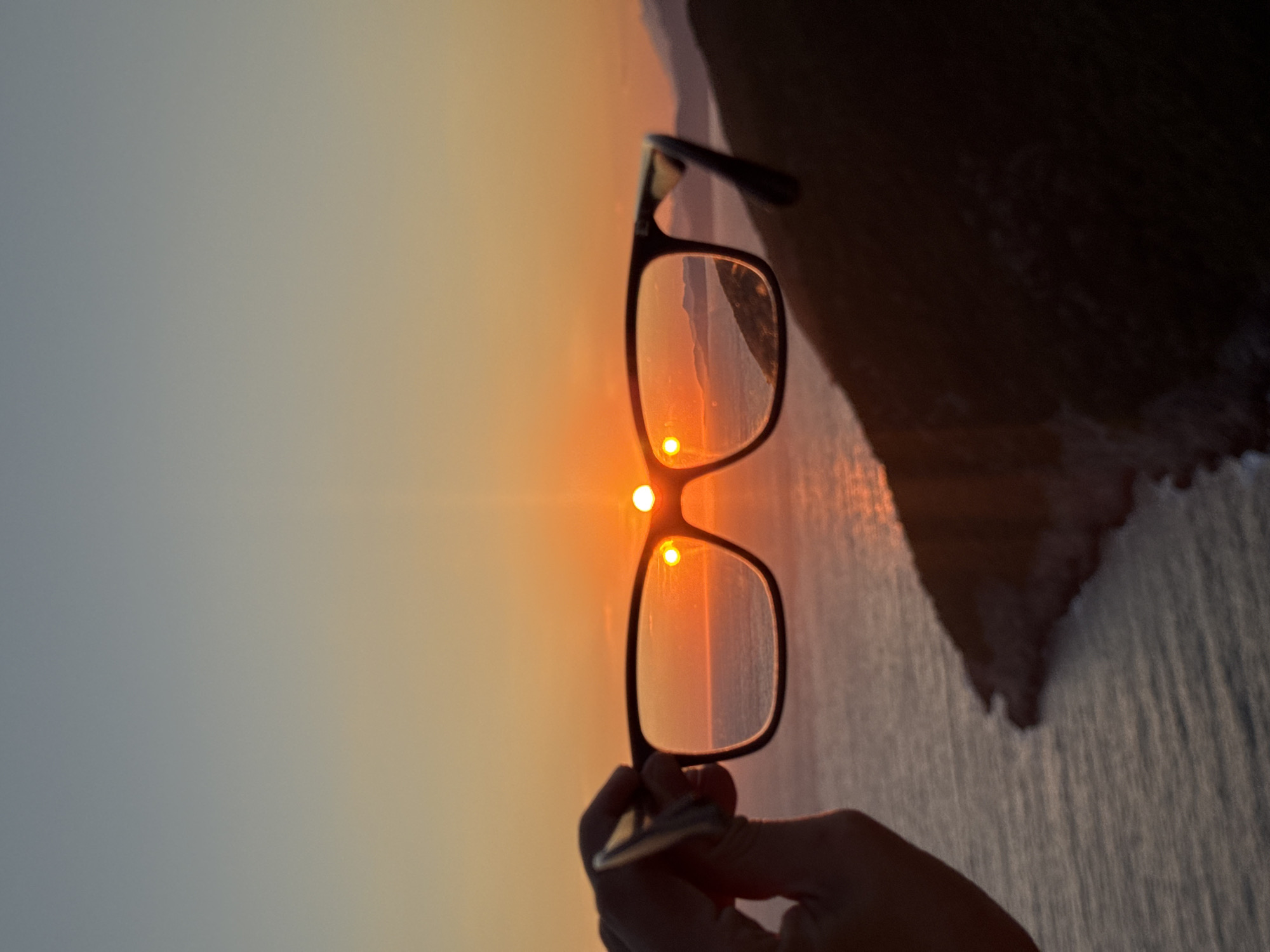 A pair of black-rimmed glasses is held in the foreground, obscuring part of a sunset over the Adriatic Sea. The sun, a bright orange orb, is partially visible through the right lens, with a smaller, reflected image of the sun appearing in the left lens. The sky transitions from a pale yellow near the horizon to a soft blue at the top. The sea below is a deep blue, with gentle waves catching the sunset's light. In the background, dark, silhouetted hills rise from the sea. The image was taken from a high vantage point, likely overlooking a coastal area. While the location is described as Zagora, Montenegro, this specific detail is not visually ascertainable from the image content itself.