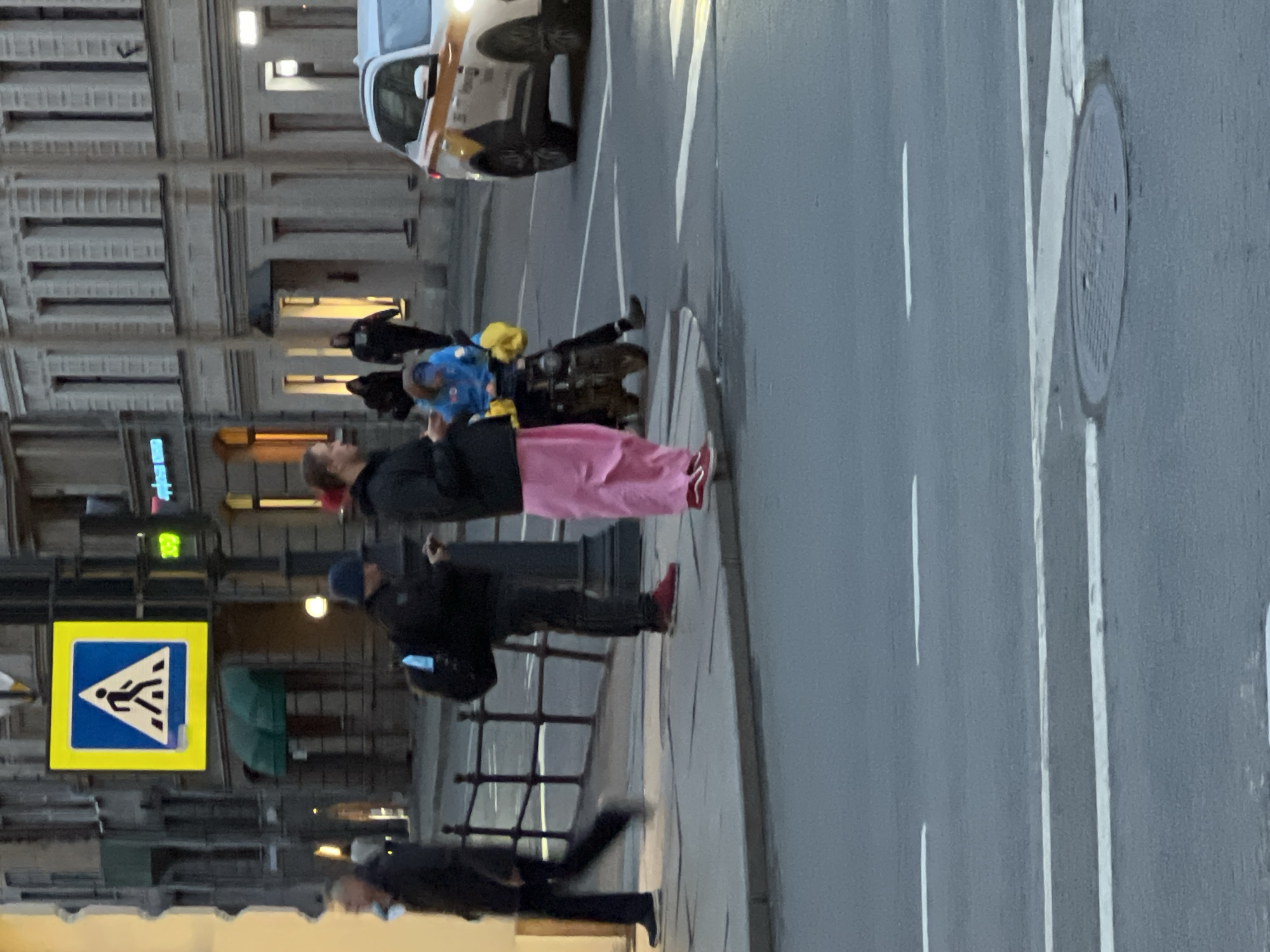 A pedestrian crosswalk in Saint Petersburg, Russia is indicated by a yellow and blue sign on a lamppost. Pedestrians, including a woman wearing a pink skirt, are waiting to cross. A vehicle is stopped in the intersection. A manhole cover is visible in the road. Several figures are walking along the sidewalk.