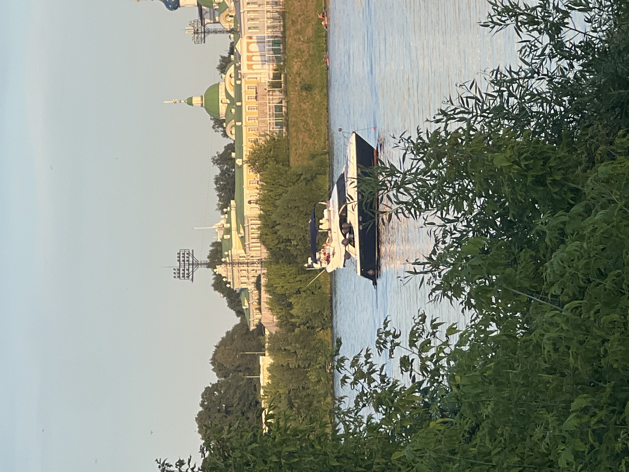 A motor yacht travels on a river in Tver, Russia. The yacht is white with a dark hull and a blue canopy. Several people are visible on the yacht. In the background, a large, pale yellow building with green domes and a tall tower is situated on a grassy embankment. Trees line the riverbank. The sky is clear and pale blue, suggesting it is either early morning or late afternoon. The water is calm with ripples. A few small boats, possibly kayaks or paddleboats, are visible further down the river.