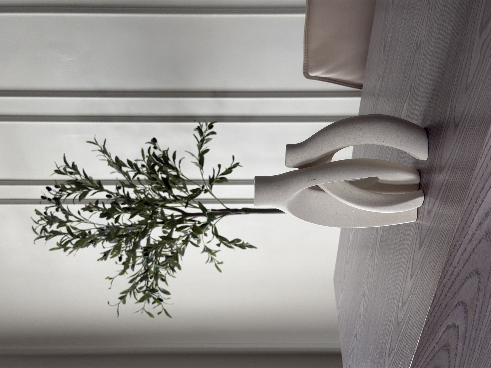 A close-up shot, possibly taken indoors, features a modern, light-colored vase with intertwining, curved elements. The vase holds a sprig of artificial olive branches with small, dark olives. The vase sits on a light-toned wooden surface with visible grain. The background is a bright, white wall with horizontal molding. A portion of a chair, upholstered in a light fabric, is visible in the upper right corner. There is no evidence suggesting this image was taken in Rostov-on-Don, Russia.