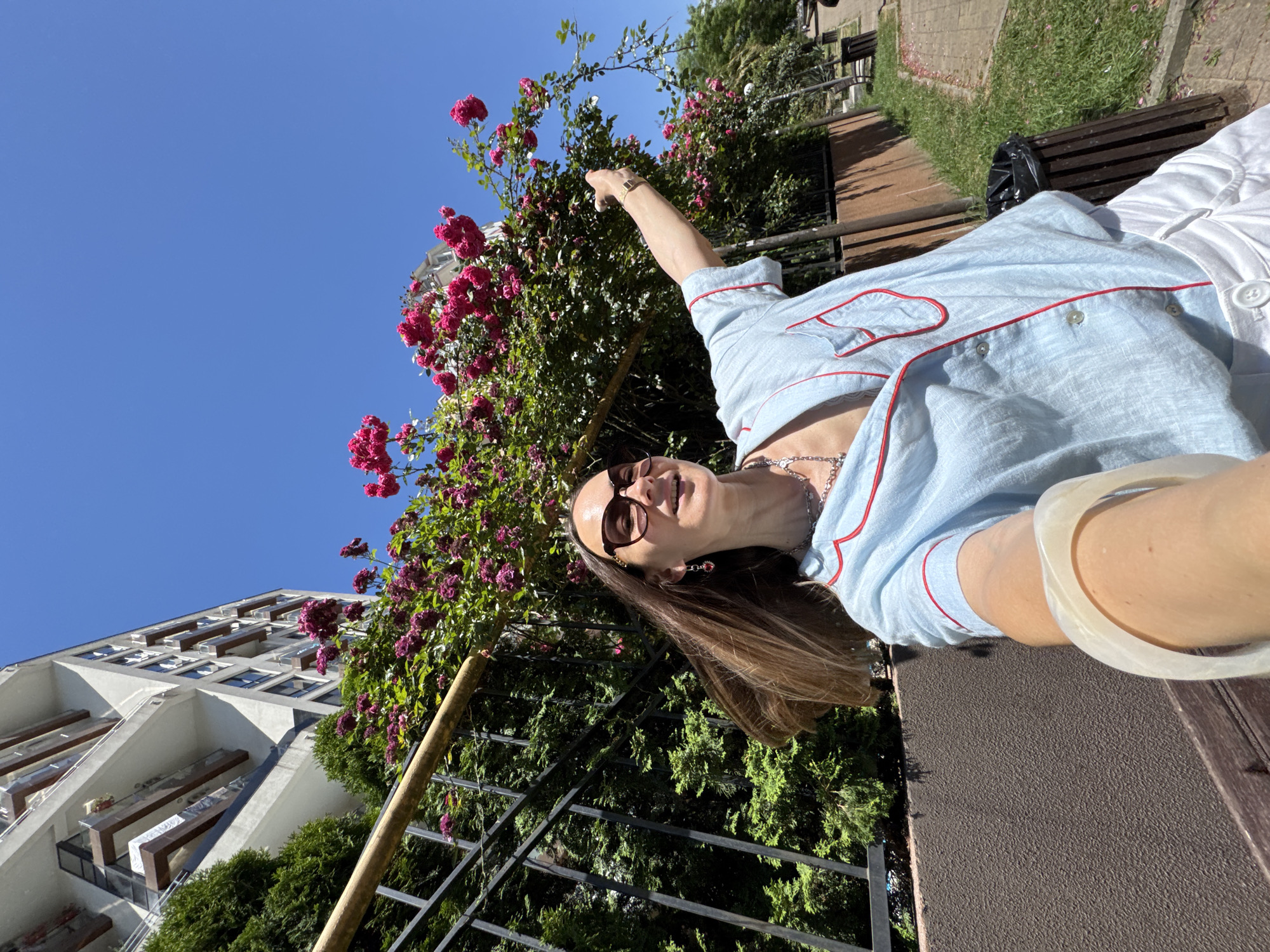 A woman, wearing sunglasses, a light blue pajama-style shirt with red trim, and white shorts, poses for a selfie outdoors. She is standing in front of a bush with bright pink flowers and a modern building with balconies. The woman is smiling and extending her right arm, pointing towards the flowers. The background includes a black metal fence, green bushes, a sidewalk, and a trash can. The image is taken on a sunny day with a clear blue sky. This scene is located in Sochi, Russia.