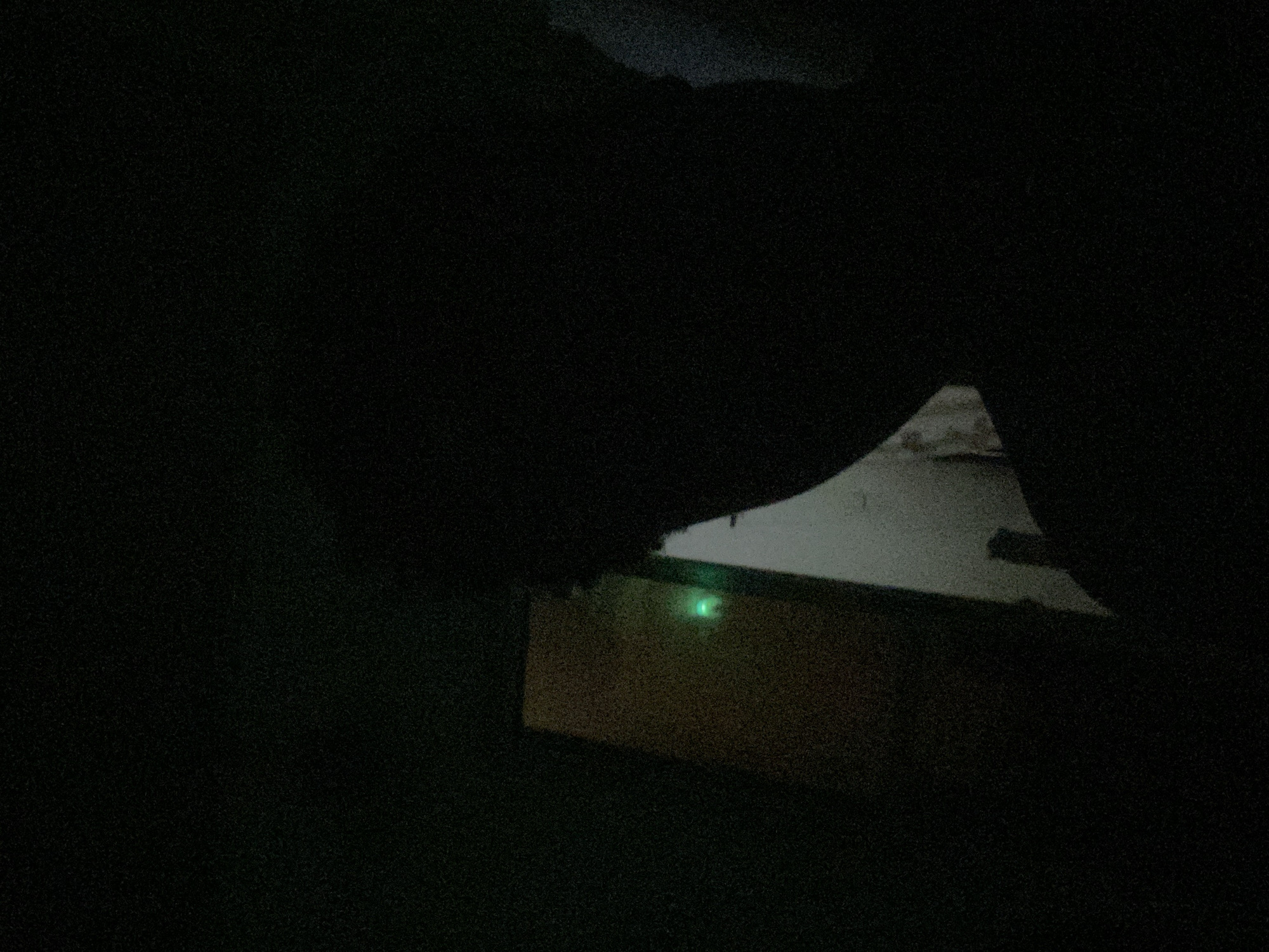 The image shows a poorly lit, dark room. A wooden structure or box with a green LED light is visible. Outside, a snowy ground is dimly illuminated. The location and presence of people and specific objects cannot be determined from the image.