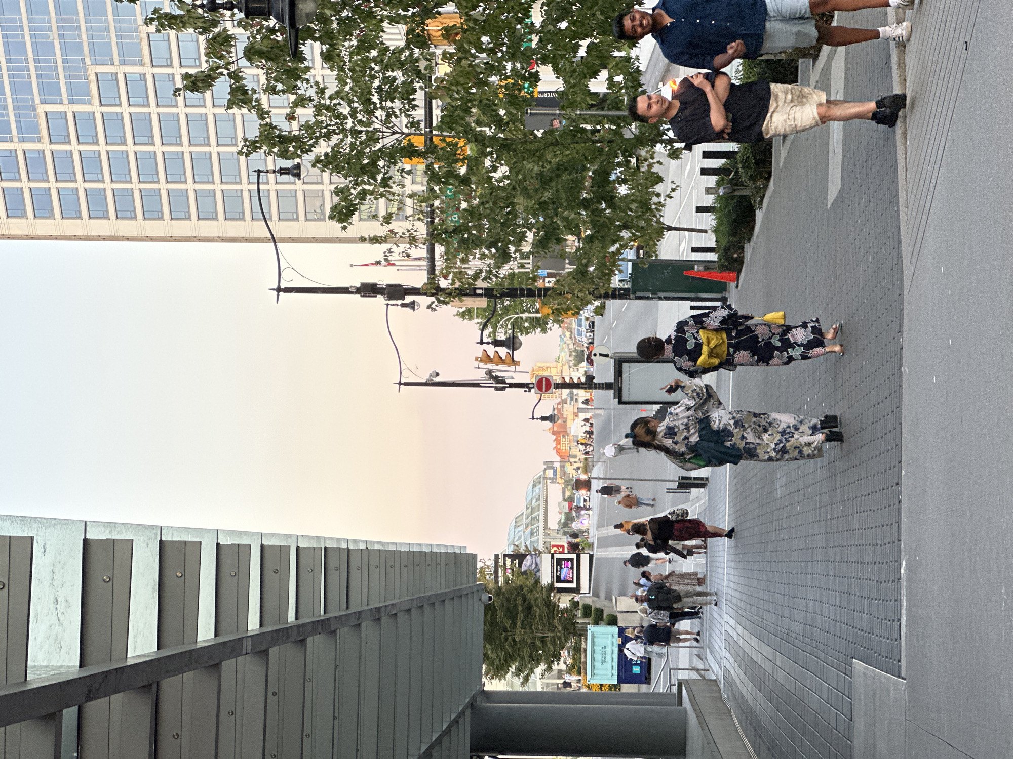 A street scene in an urban environment. In the foreground, a wide, paved walkway runs parallel to a street. On the left, a portion of a modern, covered structure with metallic overhangs is visible. Several individuals are walking on the walkway, with two women in the middle distance wearing kimonos. One kimono is floral with a blue and white pattern, and the other is dark blue with a yellow obi and floral print. A yellow purse hangs from the shoulder of the woman in the dark blue kimono. On the right side of the walkway, three men are walking. Two men in the far right are casually dressed in shorts and t-shirts. The third man, closer to the center, is wearing a black t-shirt and light-colored shorts. In the background, a tall, modern building with a grid of windows dominates the skyline, partially obscured by trees. A street with cars and streetlights extends into the distance, with more buildings and structures visible. The sky is a pale, hazy orange, suggesting either dawn or dusk.
