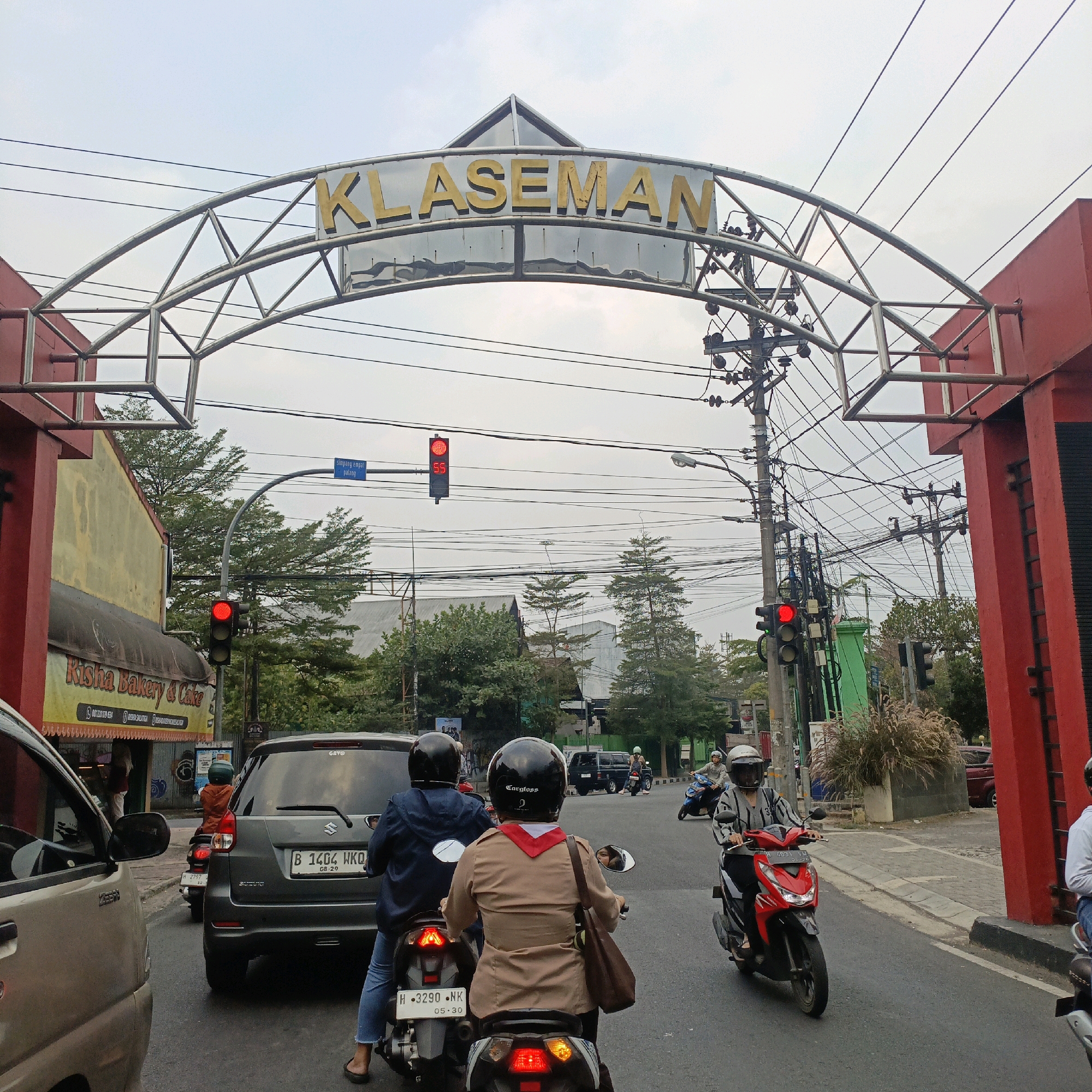 The image shows an archway with the word "KLASEMAN" in gold letters, situated over a street in Salatiga, Indonesia. Red pillars support the arch. Traffic lights are visible, with one showing red. Several vehicles are on the road, including cars and motorcycles, with people riding on them. Some individuals are wearing helmets. A banner for "Risha Bakery & Cake" is displayed on a building to the left. The sky is overcast.