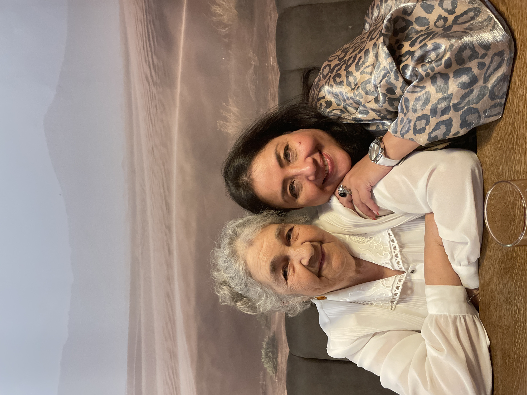 Two women are closely posed together indoors. One woman, with dark hair and wearing a leopard print top and a white watch, embraces the other woman, who has gray hair and is wearing a white lace-trimmed blouse. They are both smiling. A clear glass sits on a wooden table near the women. Behind them is a textured wall covering or backdrop depicting a desert landscape. It is not possible to determine if this photo was taken in Izhevsk, Russia.