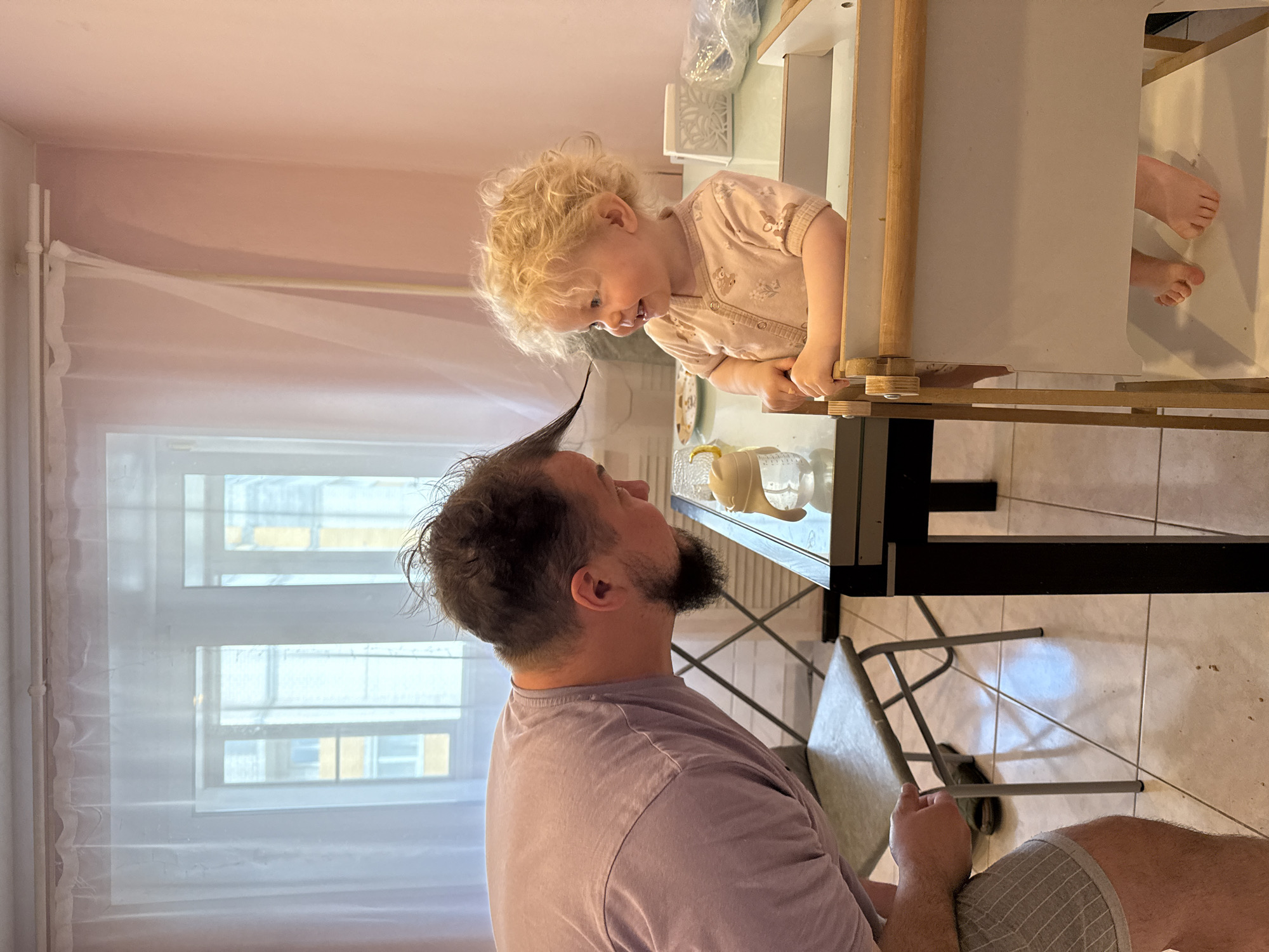 A bearded man wearing a purple t-shirt and grey patterned shorts looks up at a blonde-haired toddler who is leaning over the side of a wooden playhouse or table. The man has a tuft of hair standing straight up. The child is wearing a light-colored shirt. The scene is indoors, with a window visible to the left, covered with a sheer white curtain. Outside the window, a building can be seen. A metal-legged chair sits near the man. The playhouse or table casts a shadow on the floor. The location could be Vladimir, Russia.