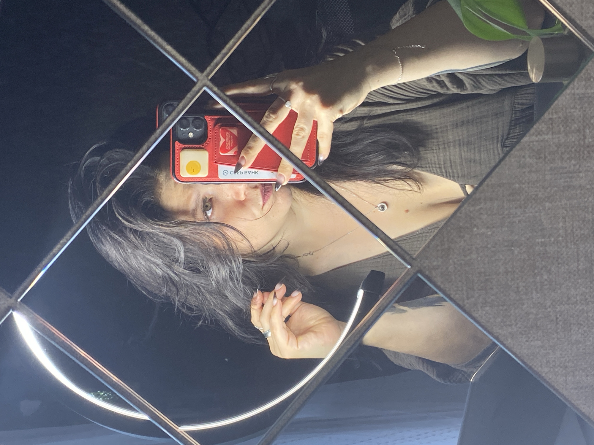 A young woman with dark, wavy hair and light brown eyes is taking a selfie in a mirror. She is wearing a brown, low-cut top and several pieces of jewelry, including rings, a necklace with a pendant, and a delicate bracelet. Her phone, with a red case featuring a yellow smiley face sticker and a heart sticker, is visible as she holds it up to take the photo. The background is dark, with a circular light source reflected in the mirror. The mirror itself is framed by square, reflective panels. There is also a glimpse of a green plant on the right side of the image. The setting is indoors, likely a personal space, and there is no visible indication of the city of Chaikovsky, Russia.