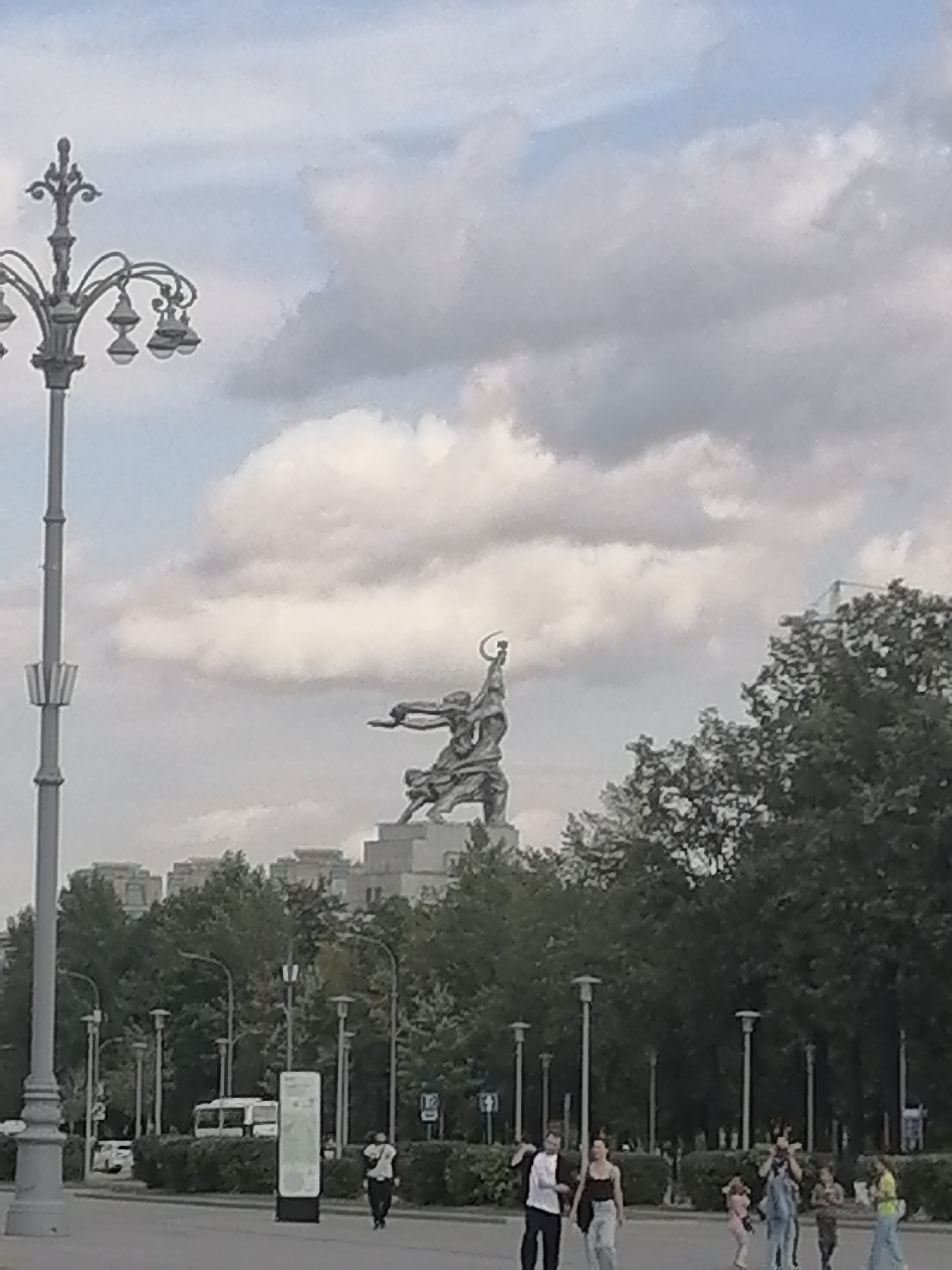 A large bronze sculpture of a man and a woman, identified as "Worker and Kolkhoz Woman" by Vera Mukhina, dominates the background of this overcast outdoor scene, set against a backdrop of dense green trees and a cityscape of light-colored buildings. The sculpture depicts the man raising a hammer and the woman holding a sickle overhead, with their figures appearing in dynamic motion. In the foreground, a paved walkway is visible with several people walking, including a man in a black shirt and pants, a woman in a black top and light-colored pants, and a group of younger individuals. A tall, ornate lamppost stands to the left of the frame, and various other streetlights are interspersed among the trees. A white bus is parked in the middle ground, and a tall, slender information kiosk is positioned near the center of the walkway. The image captures a public space in Moscow, Russia.