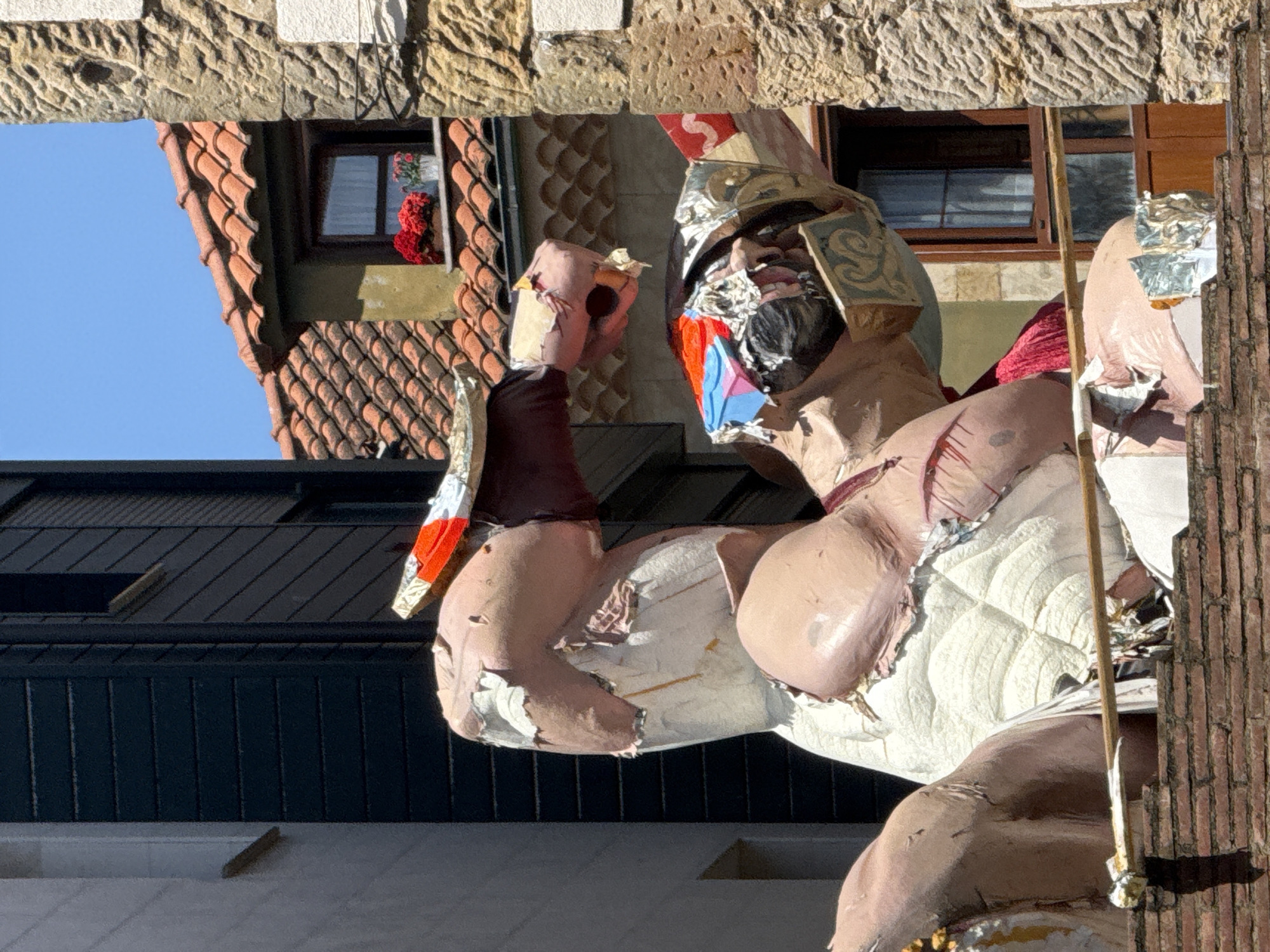 A close-up shot, framed by a stone archway, reveals a partially dilapidated, papier-mâché-style figure of a Roman gladiator. The figure displays visible decay, with peeling paint and exposed internal materials. The gladiator wears a helmet with ornate details and has painted facial features. The buildings in the background feature a red-tiled roof and a window with red flowers, while another building has a dark, modern facade. This figure is commonly seen during local festivals in Gijón/Xixón, Spain.