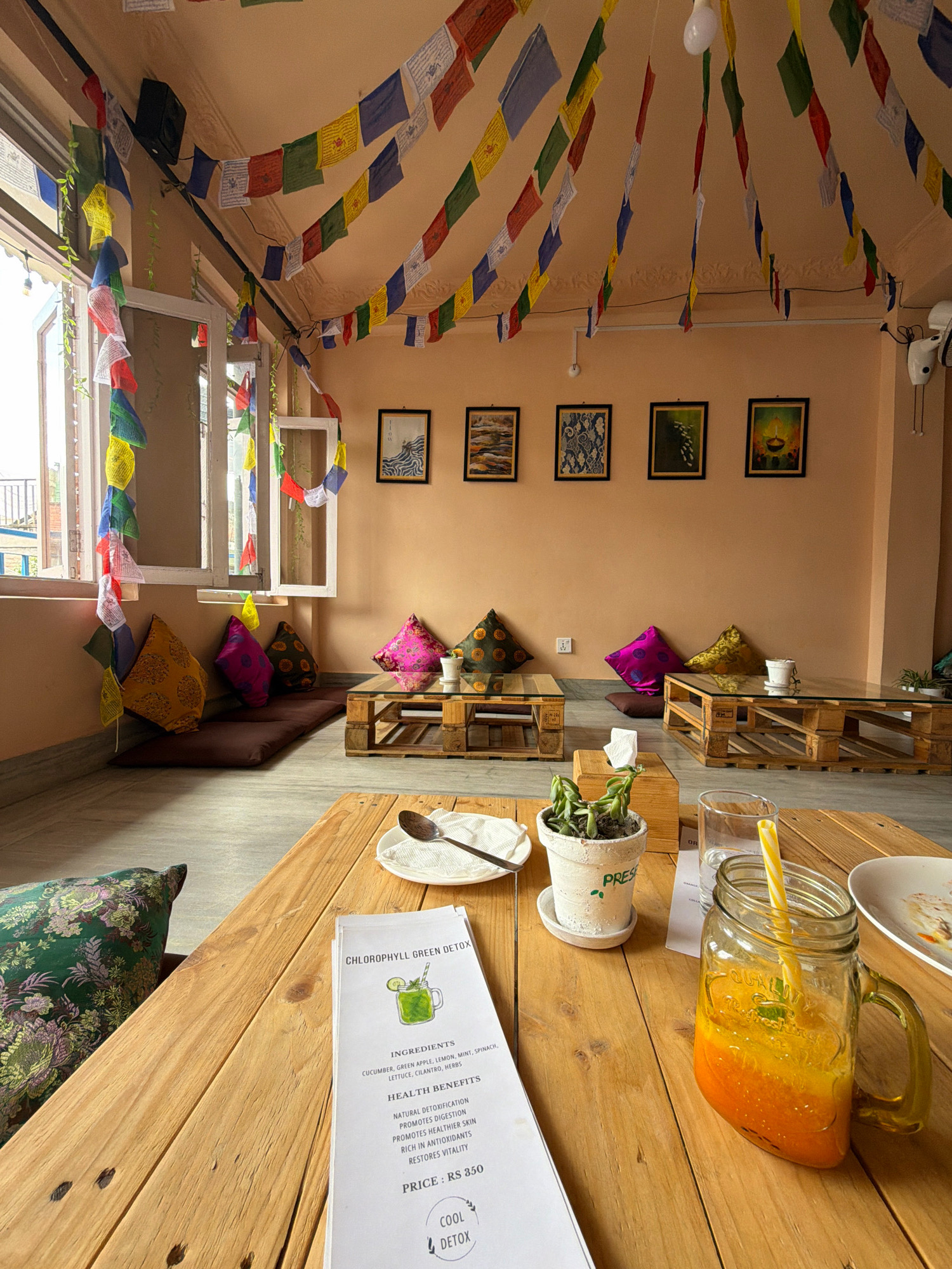 This is an interior shot of a cafe in Lalitpur, Nepal. The walls are a warm, peachy-orange color. Prayer flags are strung across the ceiling and along a window. There are several framed pieces of art on the wall. The seating consists of floor cushions and low tables made from wooden pallets. On one table, there is a mason jar with a yellow straw filled with an orange-colored drink, a glass of water, a small potted plant, and a menu. Another table has a plate with food remnants and a spoon. The overall atmosphere appears relaxed and bohemian.