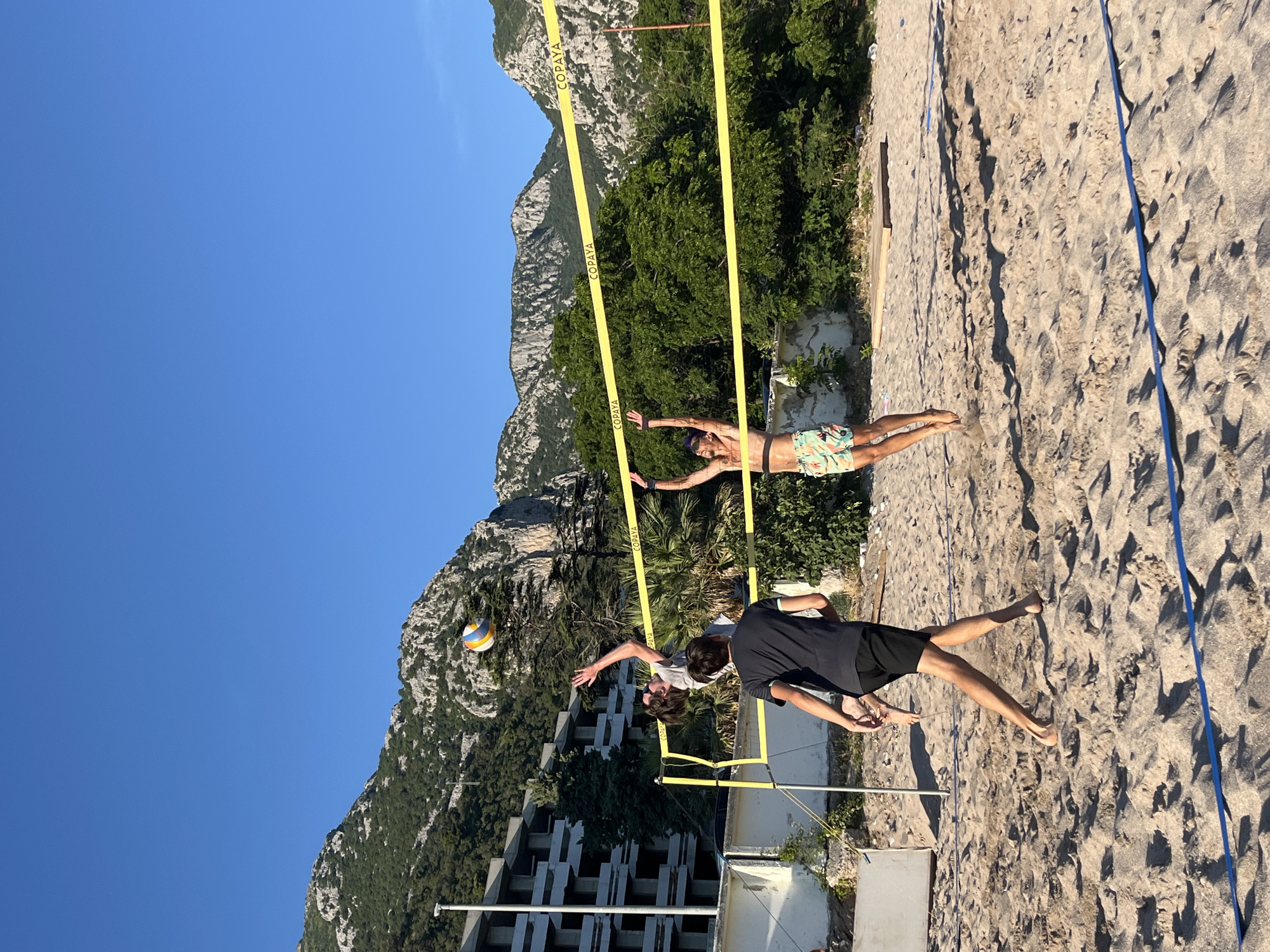 Three young men are playing beach volleyball on a sandy court. The man in the foreground, wearing a black t-shirt and black shorts, is mid-stride, his legs crossed. To his left, another man in a grey t-shirt is jumping, reaching up to hit a volleyball that is suspended in the air. To the right, a third man in patterned shorts is also jumping, his arms raised. A yellow volleyball net with "COPAYA" branding stretches across the court. In the background, a multi-story building with balconies is visible, surrounded by lush green trees and rugged, rocky mountains under a clear blue sky. The scene appears to be in Zankovići, Montenegro, a location known for its coastal setting and mountainous backdrop.