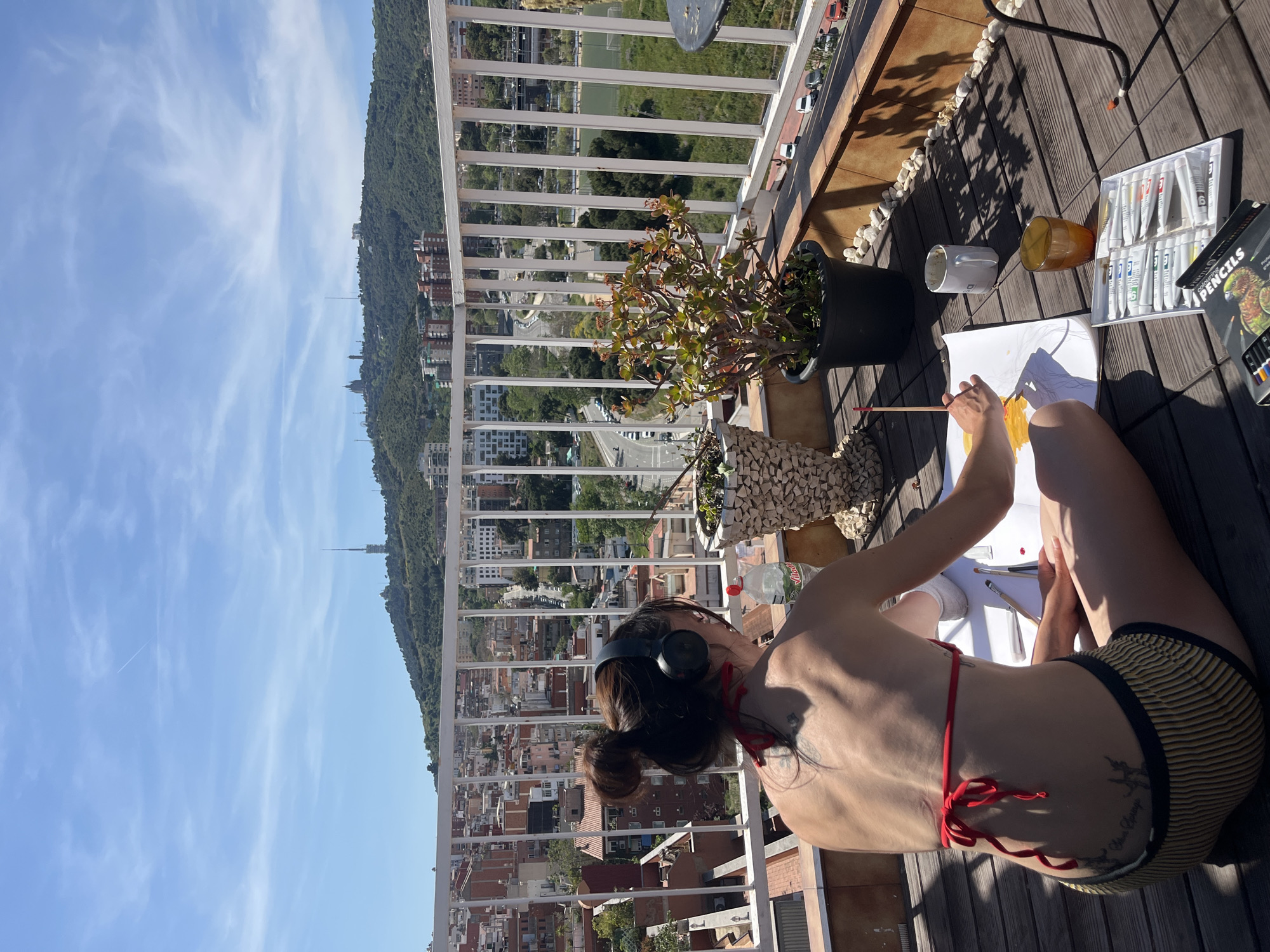 A person is painting on a balcony overlooking a city in Barcelona, Spain. The person is wearing a red bikini top, and striped yellow and black bottoms, and has headphones on. They are seated on a wooden deck with art supplies nearby. The view from the balcony includes residential buildings, a green hill, and a clear blue sky with light clouds. The balcony has a white railing and several potted plants.
