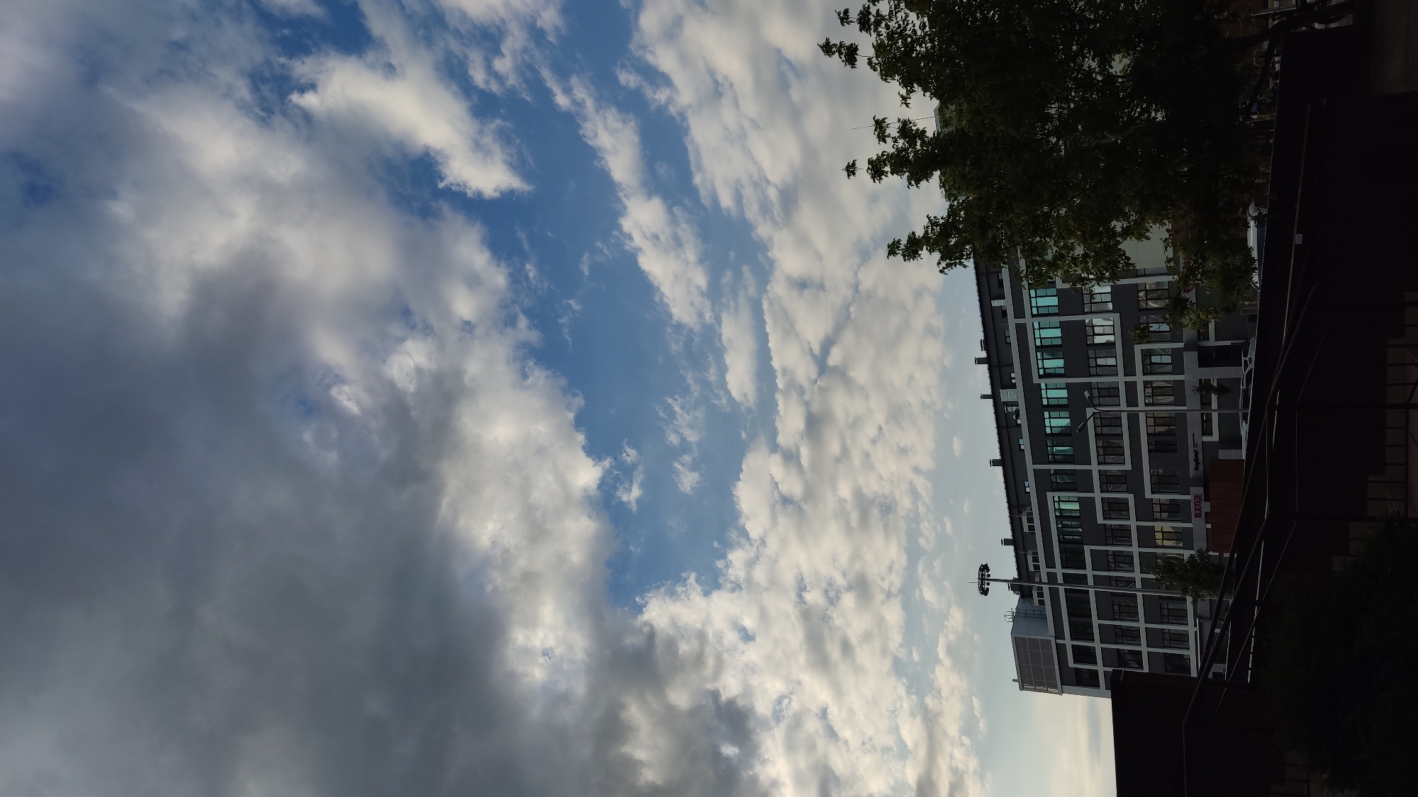 The image depicts an urban scene with a modern building and a sky filled with clouds. The building is multi-story with a dark facade and a grid-like pattern of windows, some of which reflect the blue sky. A tree with green foliage is visible in the upper right corner, partially obscuring the building. The sky is a mix of blue and white clouds, with some areas appearing darker and more overcast, suggesting either an approaching storm or the late afternoon light. The orientation of the image is vertical, with the sky occupying the left half and the building and tree on the right. There is no visible activity or people in the frame. The lighting suggests it is either early morning or late afternoon. The location is identified as Krasnodar, Russia. Some text is visible on the building, but it is too blurry to read.