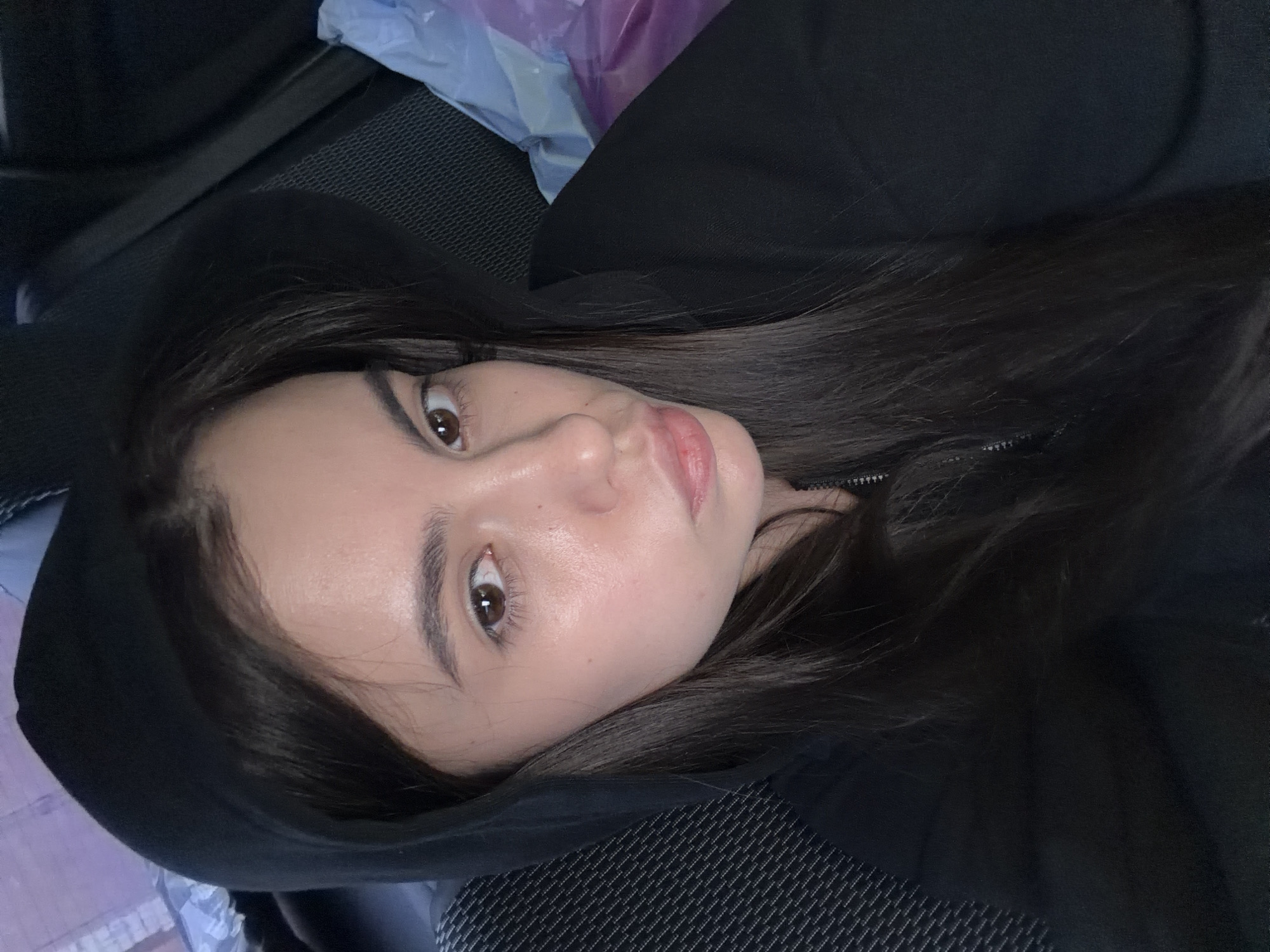 A person with long, dark hair and brown eyes is wearing a black hooded sweatshirt and looking directly at the camera. The person is in a car, with a black and gray patterned car seat visible beneath them. The background is blurred, but there appears to be a plastic bag or other material with light blue and purple colors behind the person's head. The location in the city of Chaikovsky, Russia is unknown.