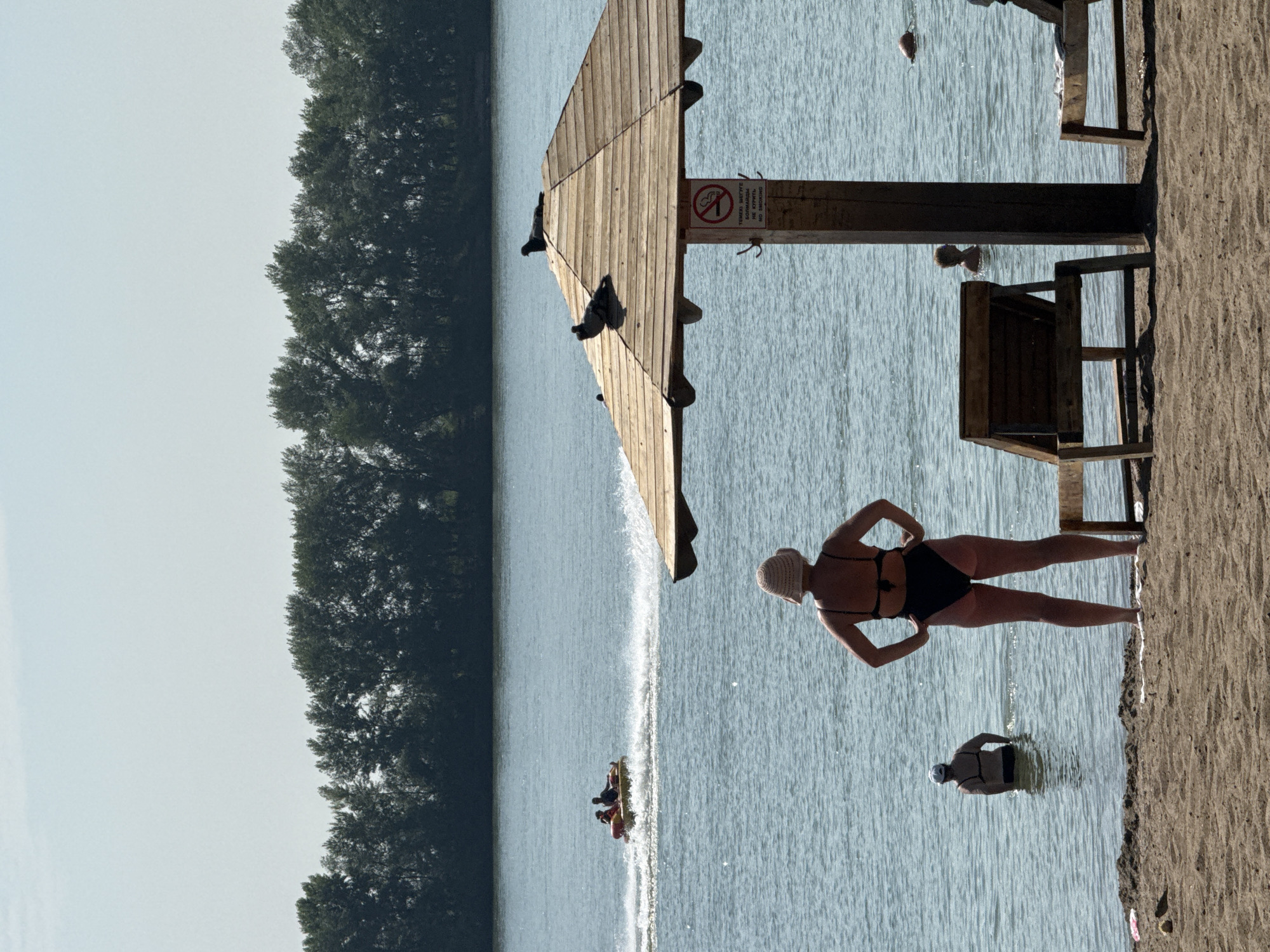 On a sunny day at the Irtysh River beach in Pavlodar, Kazakhstan, people are enjoying the water. In the foreground, a woman in a black bikini and a straw hat stands facing the water, her hands on her hips. Behind her, another woman in a black bikini and a white cap wades in the shallows. A small yellow inflatable tube is being pulled across the water in the middle ground, carrying two individuals. A wooden beach umbrella with pigeons perched on its roof shades a section of the beach. In the background, a dense line of trees runs along the riverbank under a clear sky. Two wooden lounge chairs are positioned on the sand.