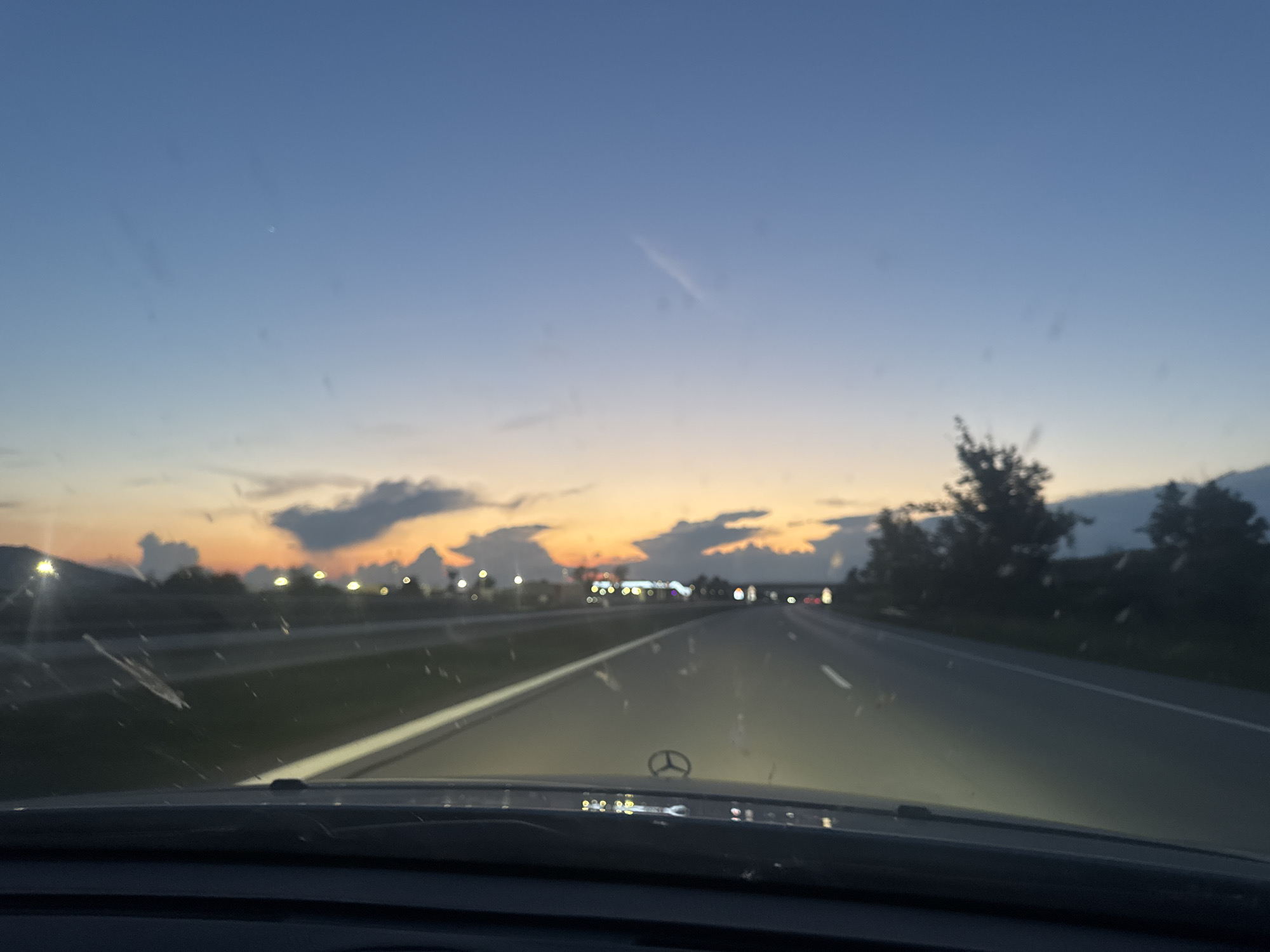 The image depicts a view from inside a vehicle driving on a highway near Enevo, Bulgaria, during sunset. The sky transitions from a deep blue at the top to vibrant orange and yellow hues near the horizon, where scattered clouds are visible. The highway stretches ahead, with multiple lanes demarcated by white lines. The headlights of the vehicle are on, illuminating the road. Distant lights, possibly from street lamps or buildings, dot the landscape along the sides of the highway. Trees and other vegetation are visible in the background. The dashboard of the vehicle, including the Mercedes-Benz logo, is in the foreground. The overall scene captures the serene atmosphere of a road trip during twilight.