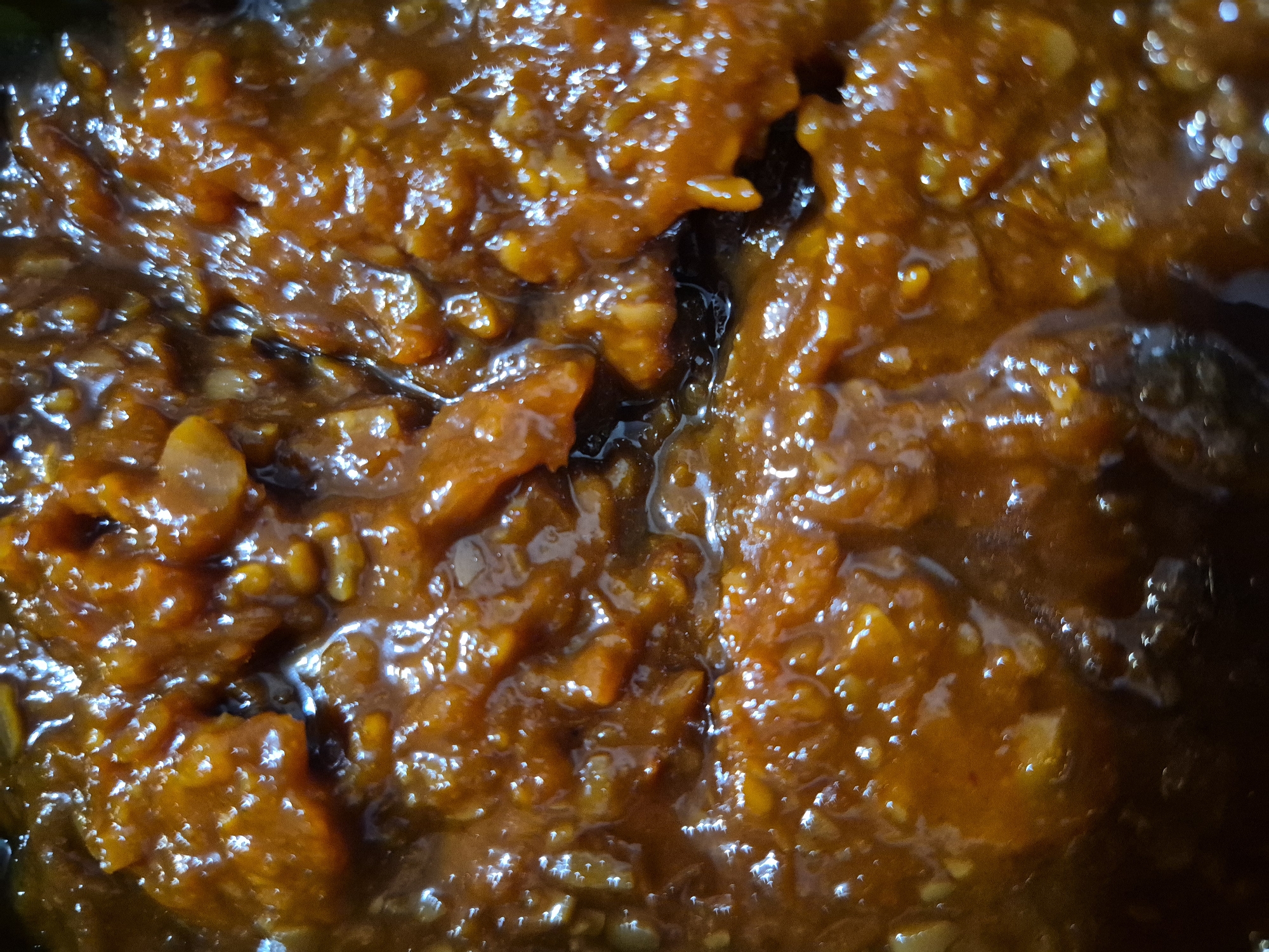 This is a close-up, overhead, macro photograph of a viscous, orange-brown, chunky sauce. The sauce appears to be slightly translucent in places, with a glossy, wet sheen that reflects the light, creating numerous bright, small, starburst-like highlights across the surface. The texture is uneven, with visible pieces of what seem to be finely diced or mashed ingredients mixed into the sauce. The overall impression is of a rich, possibly savory or sweet, condiment or prepared dish. There is no identifiable context or background that would suggest a specific location in Uglegorsk, Russia, or any other environment. The image is purely focused on the texture and appearance of the food item.