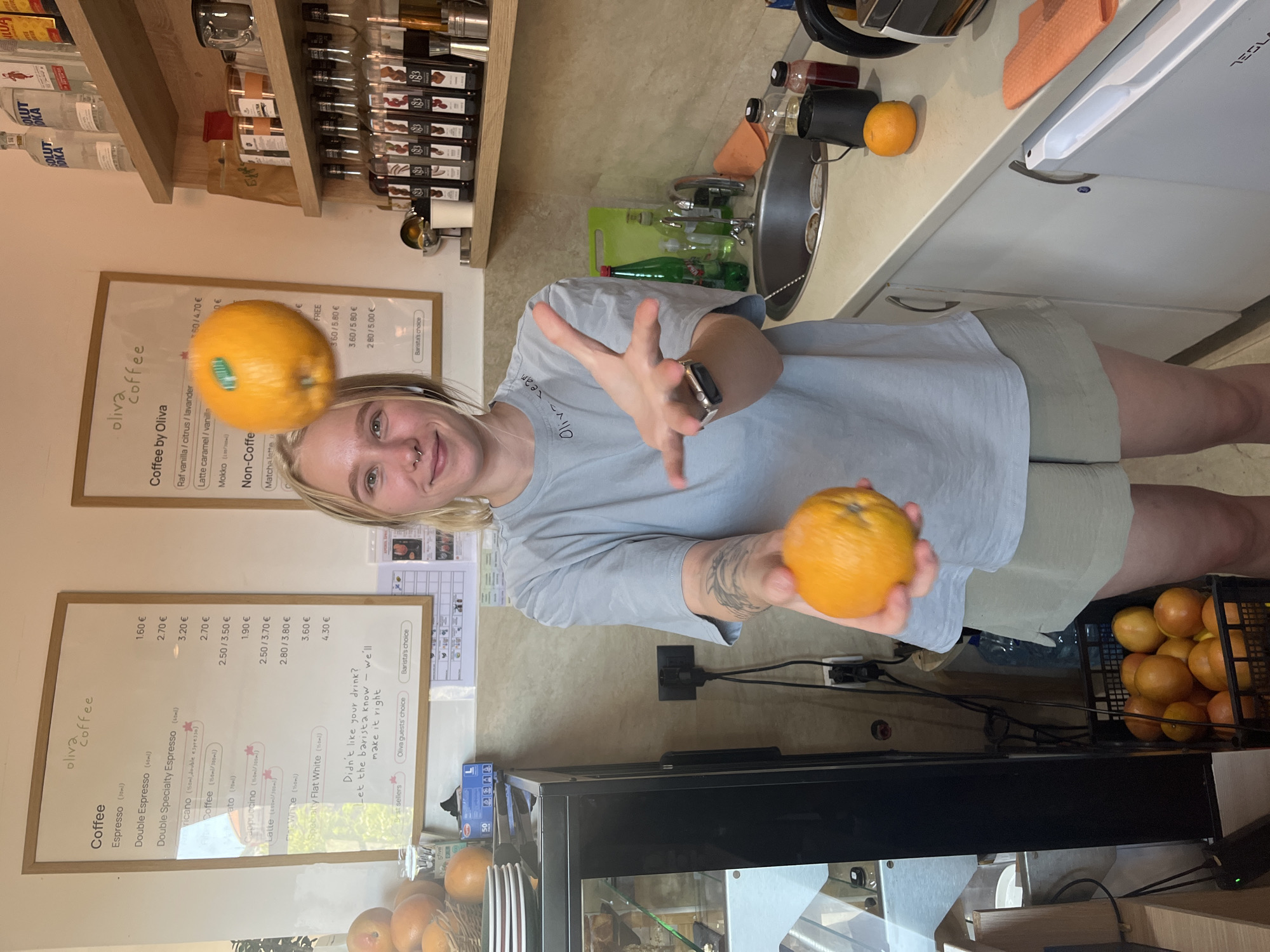 A young woman with blonde hair and nose piercings is standing in what appears to be a cafe or coffee shop. She is wearing a light blue t-shirt and beige shorts. Her right arm is raised, and she is holding an orange in her hand. Another orange is in mid-air above her head, seemingly being juggled. In the background, there are menus on the wall, shelves with bottles and cups, a coffee machine, a sink, and a basket of oranges. The scene is well-lit, suggesting an indoor environment. The cafe is likely located in Budva, Montenegro, based on the presence of what looks like a coffee menu in Montenegrin.