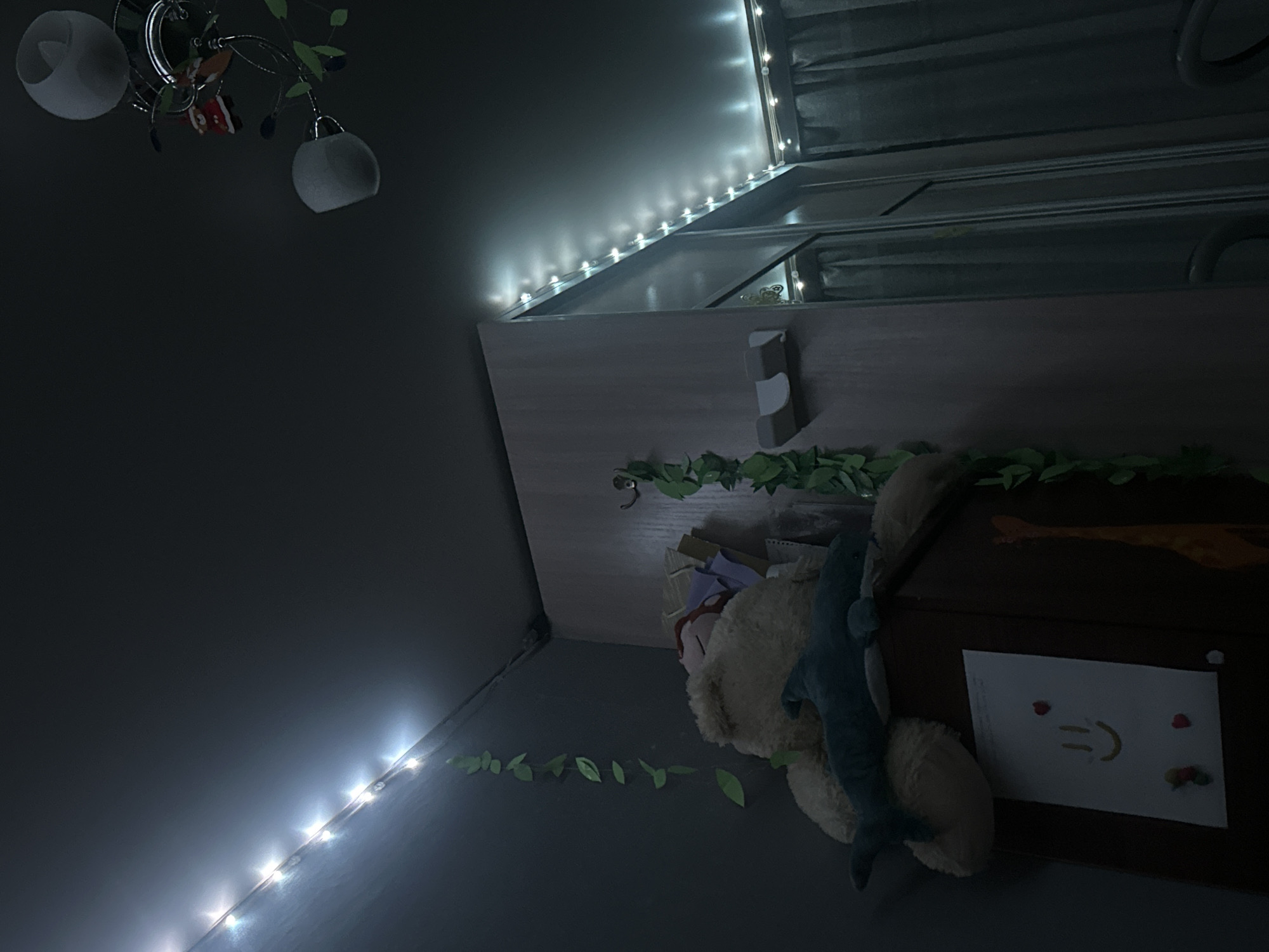 The image shows a dimly lit room, possibly a child's bedroom, in Petropavlovsk-Kamchatsky, Russia. Decorative string lights with white LEDs are illuminating the ceiling and the upper parts of the walls. On a shelf, a large stuffed teddy bear is visible, with a plush shark lying on top of it. To the left of the teddy bear, a string of artificial green leaves hangs down the wall. A dark brown cabinet is below the shelf, with a drawing of a smiley face and two red heart shapes attached to its front. To the right, a large wardrobe or closet with mirrored doors is present, and gray curtains hang behind it. A ceiling light fixture with two white lampshades and some decorative elements is also visible.