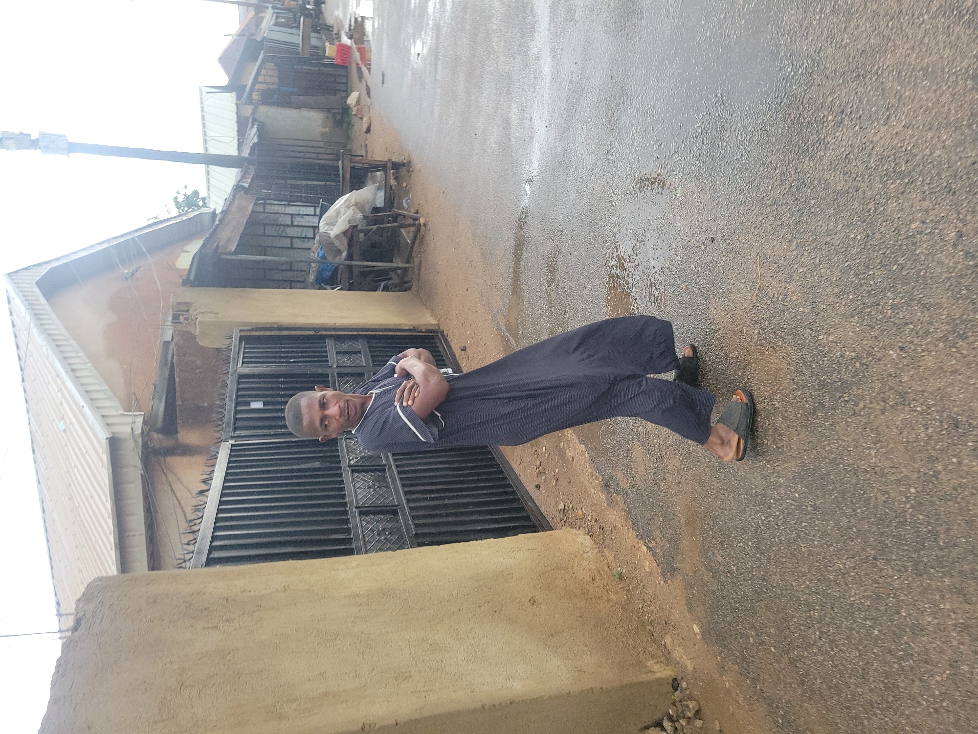 A young man stands with his arms crossed on a wet, paved road in what appears to be an urban area. He is wearing a dark, floor-length robe with white trim. The man is looking to his right and slightly away from the camera. Behind him, a dark metal gate with pointed spikes is visible, in front of a building with a corrugated metal roof. The ground is wet, with several puddles reflecting the overcast sky. In the background, a series of stalls or shops with similar roofing are visible, some with covered goods. A cell tower is visible in the distance. The scene suggests a residential or commercial street in Jos, Nigeria, likely after rain.