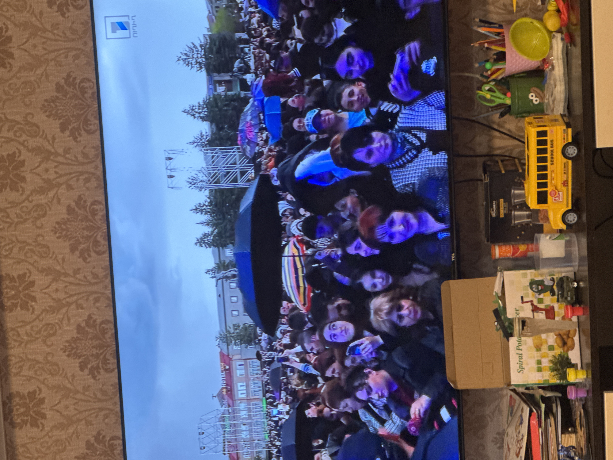 On a television screen, a dense crowd is visible, likely gathered in a square in Yerevan, Armenia. The square is lined with buildings, including what appears to be a hotel. Trees and a stage structure are visible. The crowd consists of many individuals, some holding phones, others with umbrellas. A variety of colors, especially blues and purples, illuminate the crowd. To the right of the screen, on what is likely a side table, are various items including school supplies, a toy school bus, and a box labeled "Spiral Potato." The screen is situated against a backdrop of patterned wallpaper.