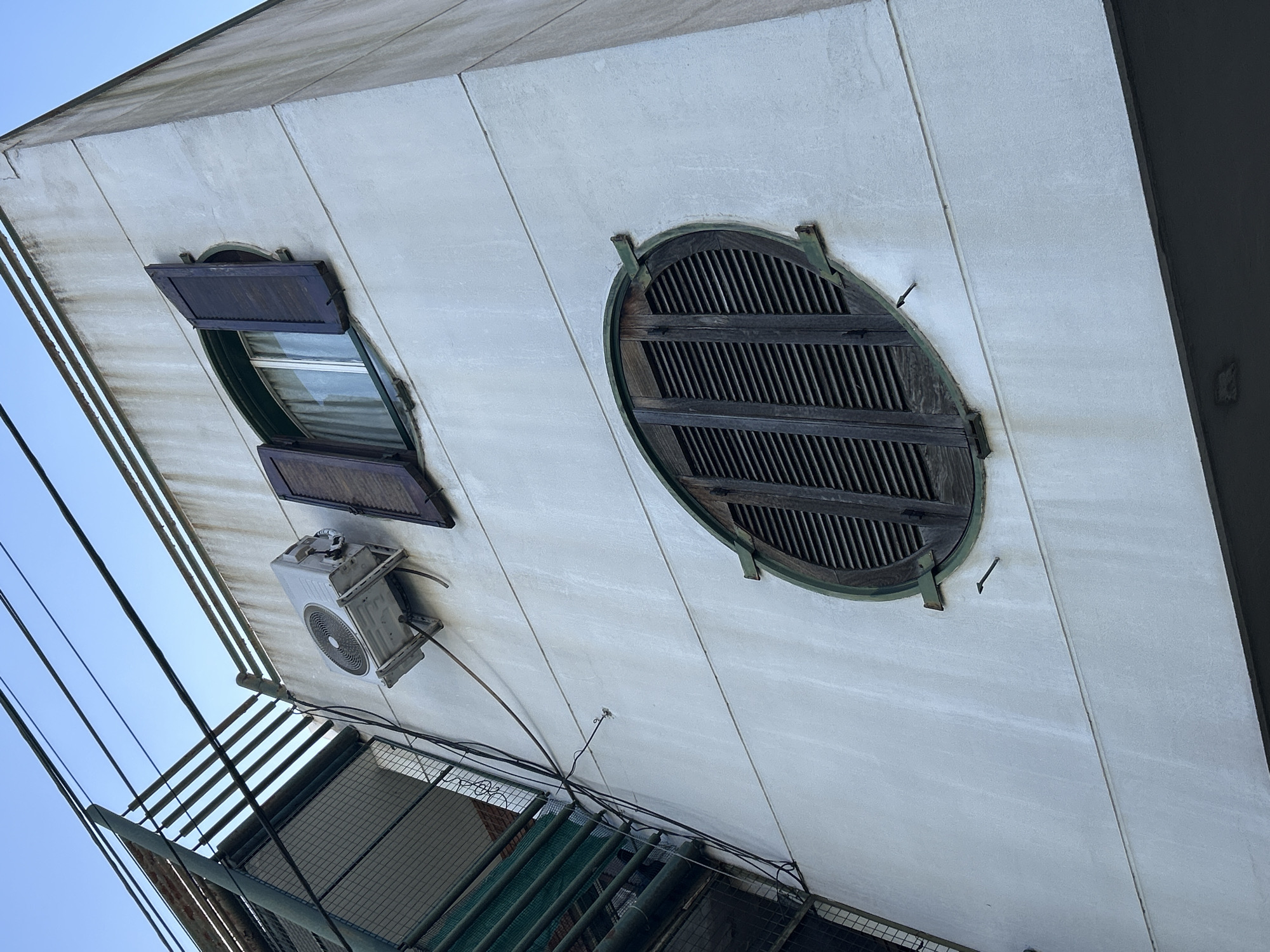 A low-angle shot captures the exterior of a pale, aged building in Buenos Aires, Argentina, under a clear blue sky. On the upper portion of the wall, a white air conditioning unit is mounted, angled downwards. To its right, a rectangular window features dark wooden shutters, one of which is open. Below this, a distinctive oval window is covered by closed dark wooden louvre shutters, framed by a green trim. Electrical wires crisscross the sky above the building, and a section of a metallic structure, possibly a balcony or fire escape, is visible on the left. The building's facade exhibits signs of wear, with staining and discoloration.