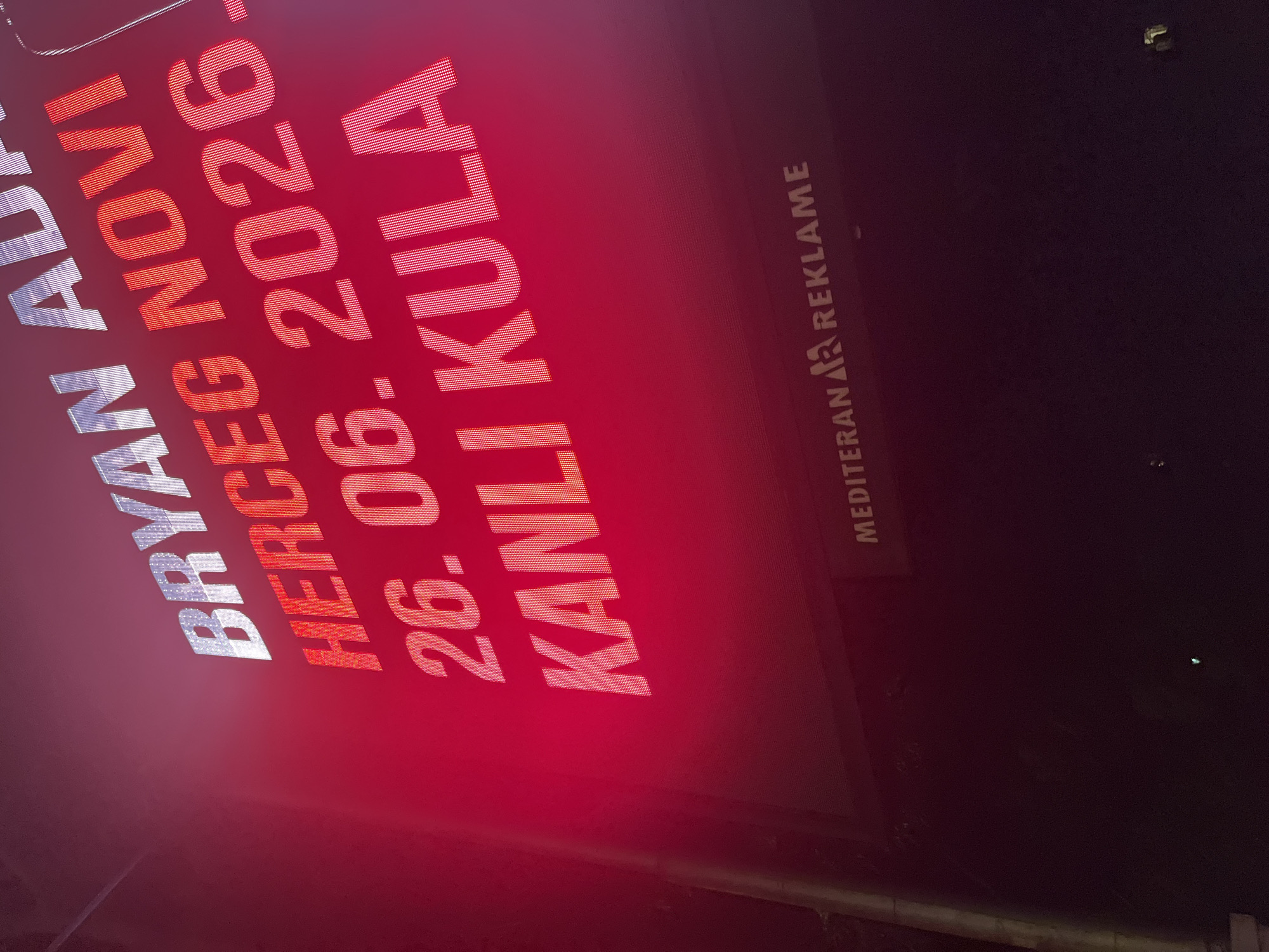 The image shows a large digital billboard in Budva, Montenegro, displaying text in white and red. The top line of text reads "BRYAN AD". Below that, in red, is "HERCEG NOVI". The next line, also in red, reads "26.06.2026 -". The final line of red text is "KANLI KULA". Towards the bottom of the billboard, smaller white text reads "MEDITERAN REKLAME". The billboard is illuminated and appears to be outdoors at night, with the surrounding environment mostly dark. The red glow from the billboard dominates the image.