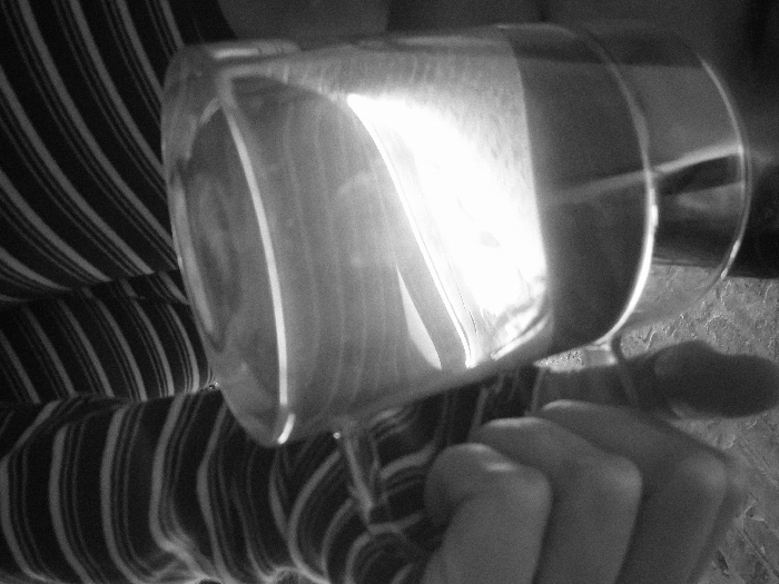A black and white image features a hand gripping a drinking glass. The glass is positioned horizontally with the opening facing the viewer. A striped garment is visible behind the glass. Light reflects intensely off the glass surface, obscuring the contents. The hand appears to be lightly flexed, holding the glass securely. The surrounding background is out of focus, adding emphasis to the glass and hand.