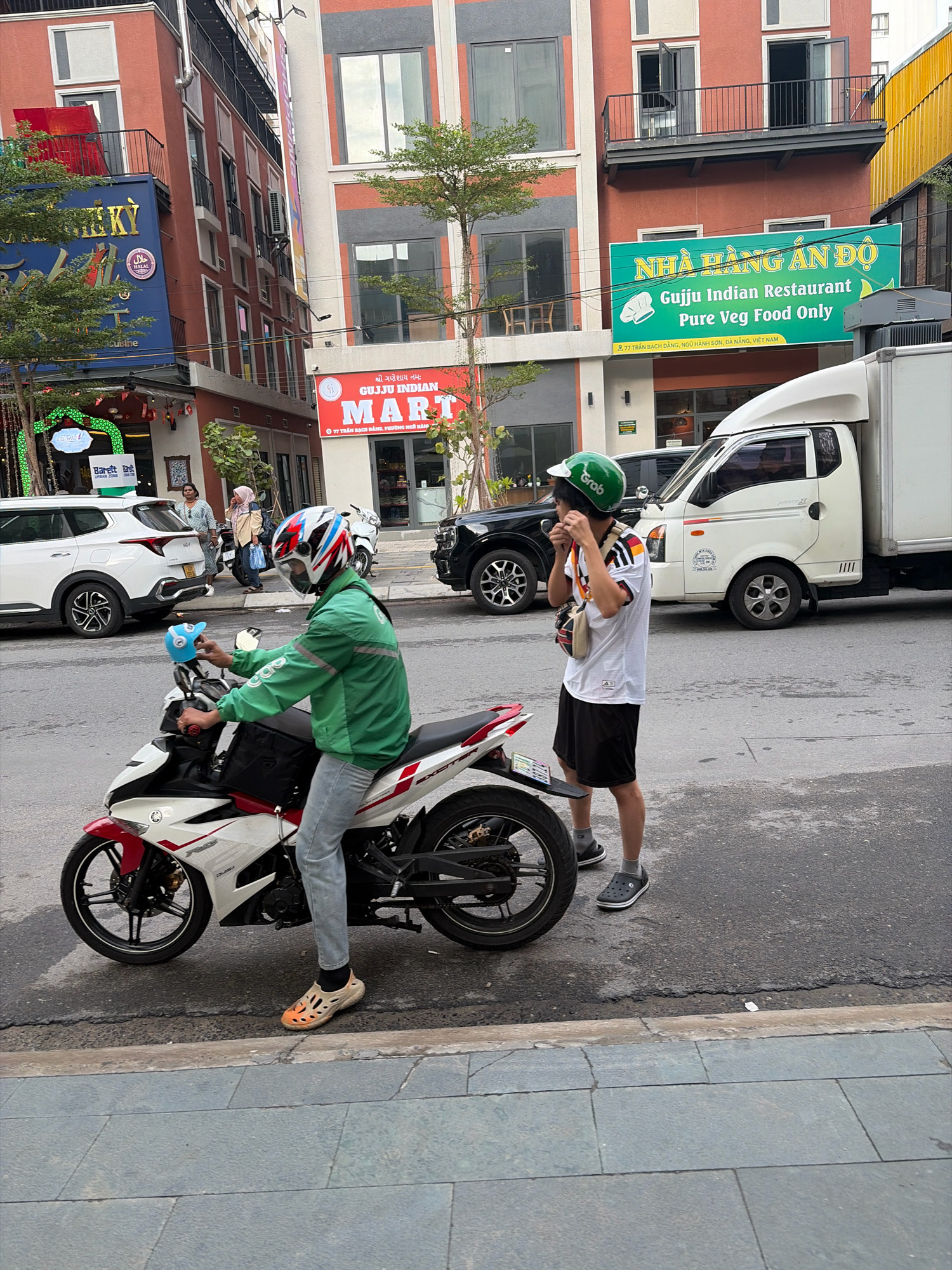 The image depicts a street scene in Da Nang, Vietnam, likely in a commercial area. In the foreground, a person wearing a green jacket, grey pants, and a helmet is seated on a white and red motorcycle. The motorcycle has a black storage box on the back. The rider is holding a blue child's helmet. To the right of the motorcycle, another person wearing a white shirt with black stripes, black shorts, and a green helmet is standing. This individual is adjusting their helmet and carrying a beige shoulder bag. Behind them, a white truck is parked. The background features buildings with storefronts, including a sign for "Guijo Indian Restaurant Pure Veg Food Only" and another for "MART". Trees and streetlights are also visible. The street is paved, and a sidewalk is present in the foreground. The overall impression is a snapshot of daily life in a Vietnamese city.