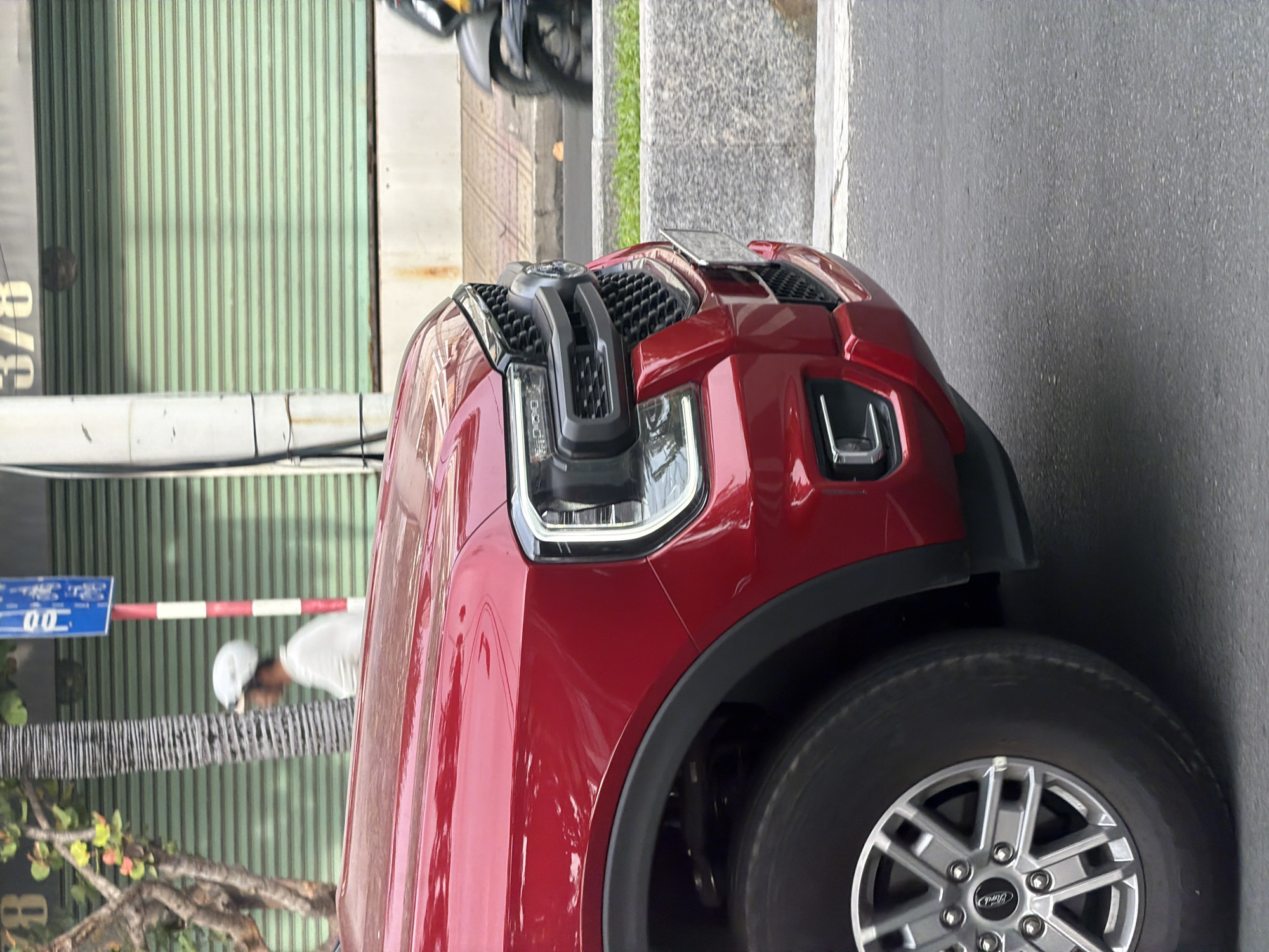 A red Ford Ranger pickup truck is parked on a city street in Nha Trang Ward, Vietnam. The front driver's side of the truck is visible, including the wheel, fender, headlight, and grille. The truck is parked on an asphalt road next to a concrete barrier and some grass. In the background, a person wearing a white helmet and a light-colored shirt is walking past a tree and a building with a green rolling door. A motorcycle is also visible in the background, to the right of the truck. The scene appears to be during daylight.