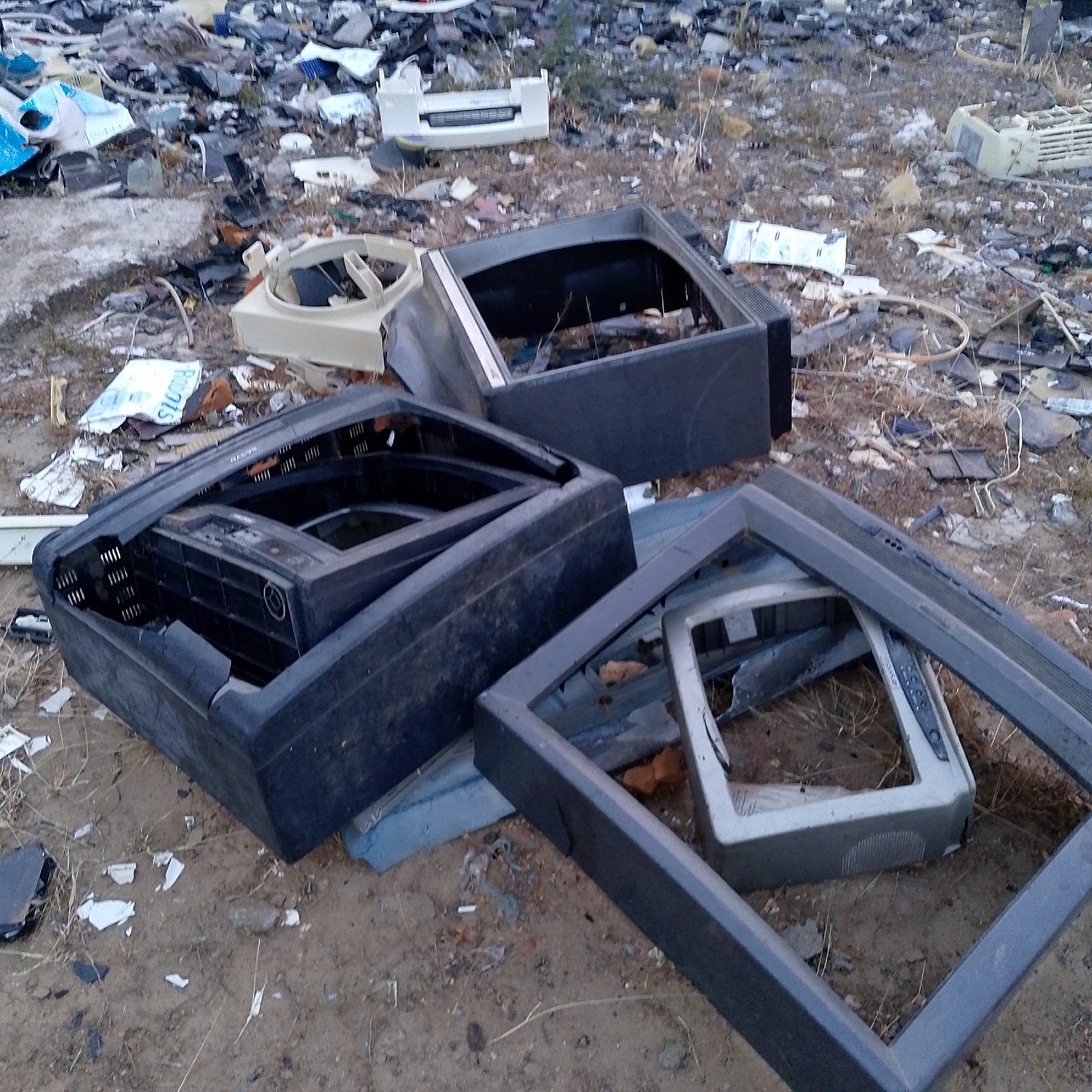 The image shows an outdoor scene in what appears to be a landfill or dumping ground. There are several discarded computer monitors and parts strewn across the ground, including monitor casings, internal components, and other electronic waste. The ground is covered in dirt, debris, and dry vegetation. There are no people visible in the image. The exact location in the city of Altagracia, Venezuela, is not identifiable from the provided image.