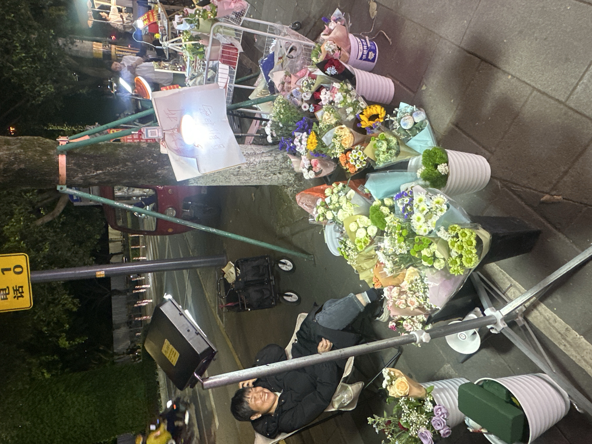 The image depicts a lively night scene of a street flower vendor in the Chongqing High-tech Zone, China. The setting is an urban sidewalk, illuminated by artificial lights, indicating it is nighttime.
In the foreground, a young man, likely the vendor, is comfortably reclined on a folding chair, wearing a dark puffer jacket and jeans. He is looking towards the camera with a cheerful, relaxed smile. Around him, an abundant display of fresh flower bouquets is arranged in white ribbed buckets and wrapped in various colored papers and plastics. The diverse selection includes white, green, yellow, orange, purple, and red blooms, featuring flowers such as roses, sunflowers, chrysanthemums, and other mixed varieties.
The flower display is set up on a metal rack. A bright light panel, possibly for illumination or attracting attention, shines intensely on a white sign, making the Chinese characters written on it difficult to decipher due to glare. Another black rectangular light fixture is visible on a stand nearby. Behind the seated vendor, a black utility cart with bags suggests mobile equipment.
In the background, another person is partially visible near what appears to be another stall or shop. A large tree trunk, supported by green metal poles, stands prominently on the left. A red vehicle, possibly a small tricycle or utility truck, is partly seen behind the tree. A yellow rectangular street sign on a pole clearly displays the Chinese characters "电话" (diànhuà, meaning "telephone") and the number "10". The ground is a dark paved surface. The overall atmosphere suggests a bustling, yet relaxed, evening market or street vending operation.